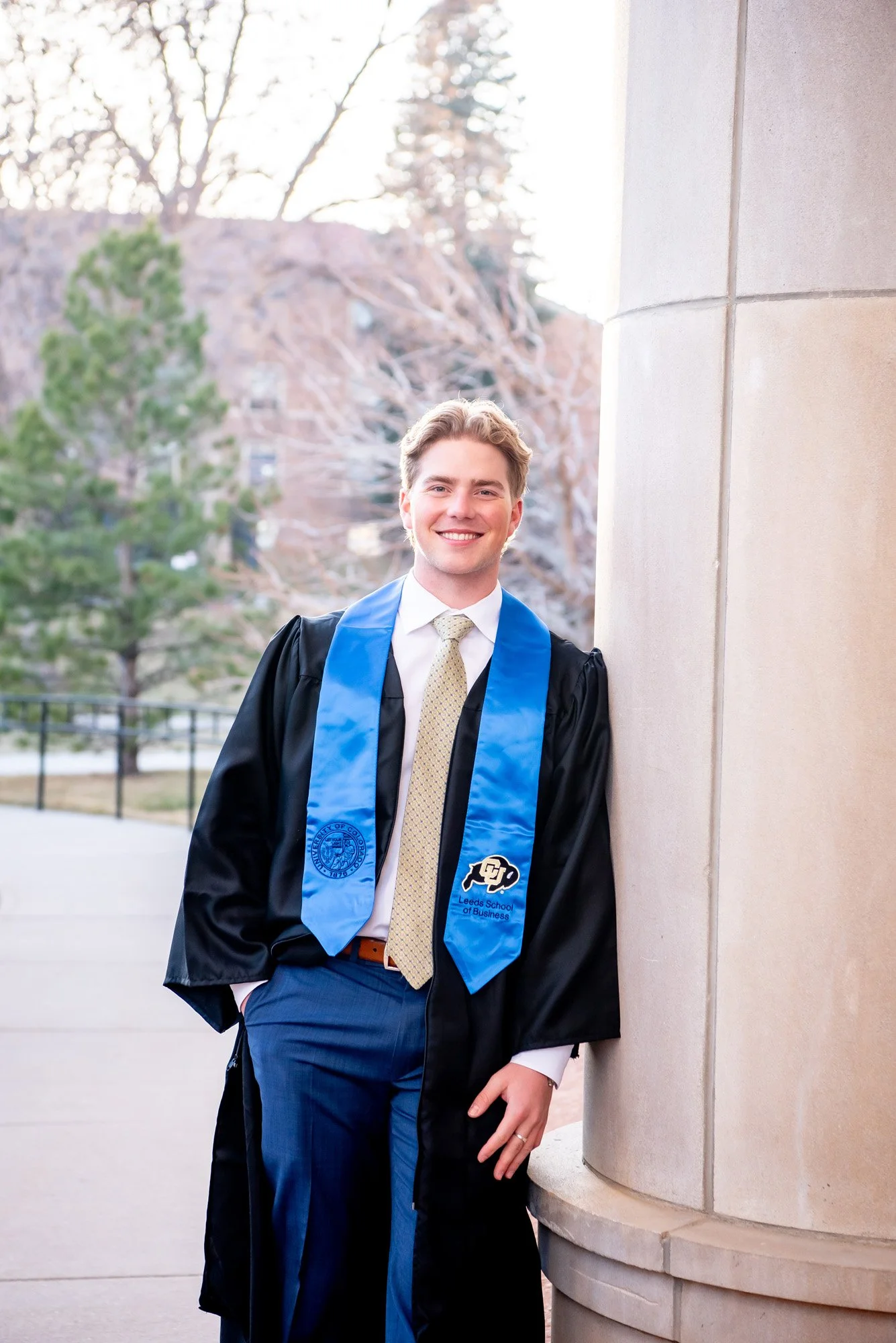 A young man in a black graduation gown and blue stole with the University of Colorado Boulder logo, smiling and leaning against a stone pillar outdoors during daytime.