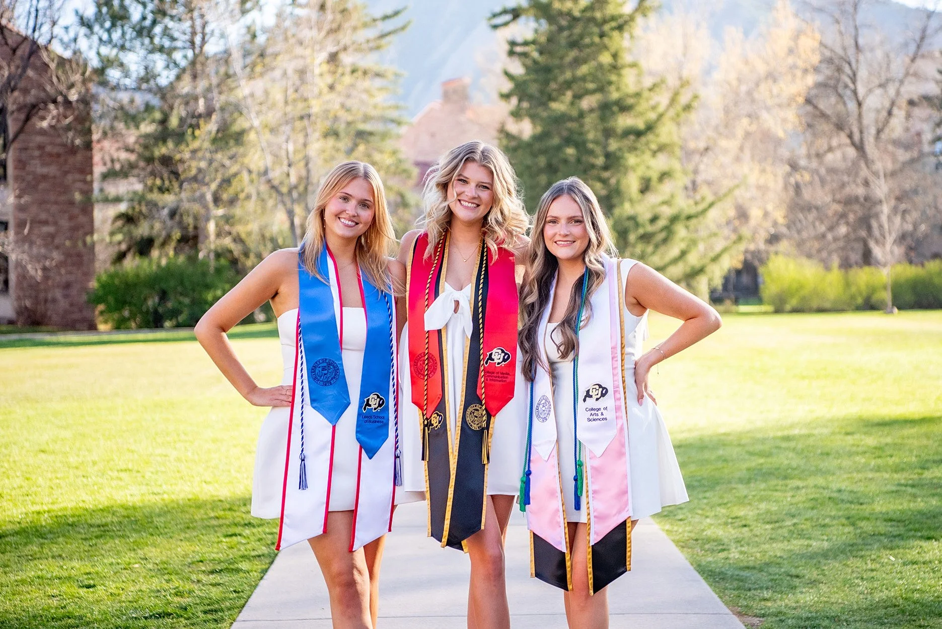 Three women at a graduation ceremony wearing caps and gowns with honor cords and stoles, standing outdoors on a sunny day with trees and buildings in the background.
