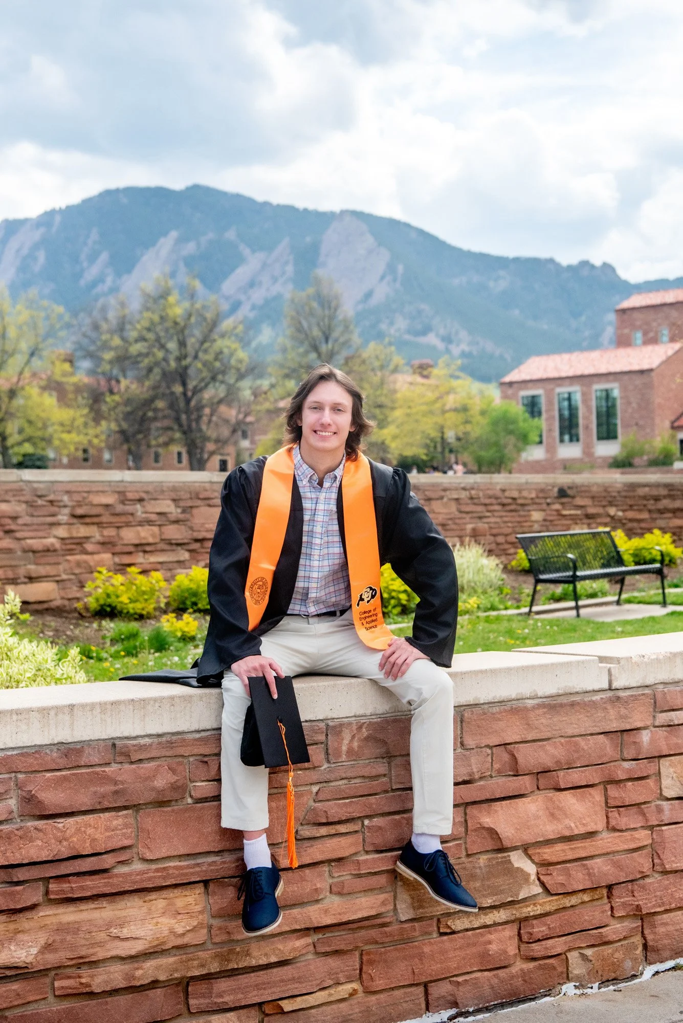 A young man in a graduation gown and cap sitting on a brick wall outdoors, smiling, with a mountainous background and buildings.