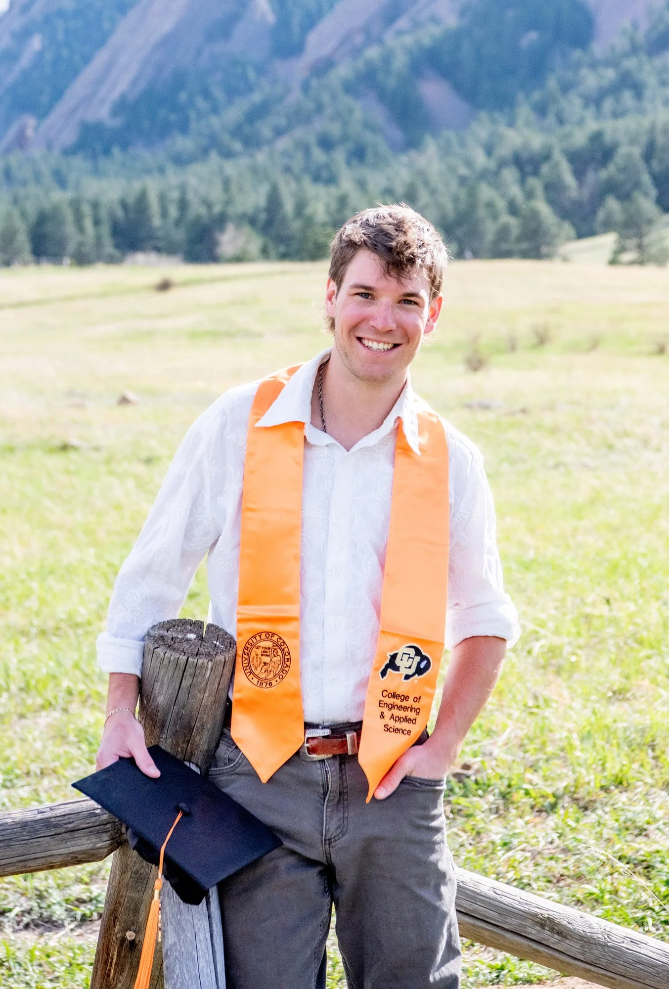 A young man in a white shirt with rolled-up sleeves, wearing a graduation stole from the University of Colorado College of Engineering & Applied Science, stands outdoors in a grassy field with mountains in the background. He is smiling and holding a graduation cap and diploma.