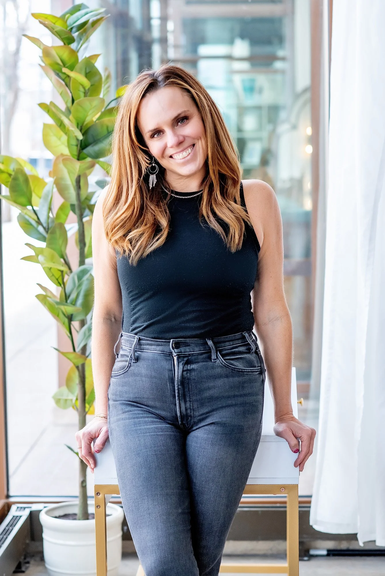 A smiling woman with long, wavy auburn hair wearing a black sleeveless top and dark jeans, standing indoors near a large window with a green potted plant in the background.