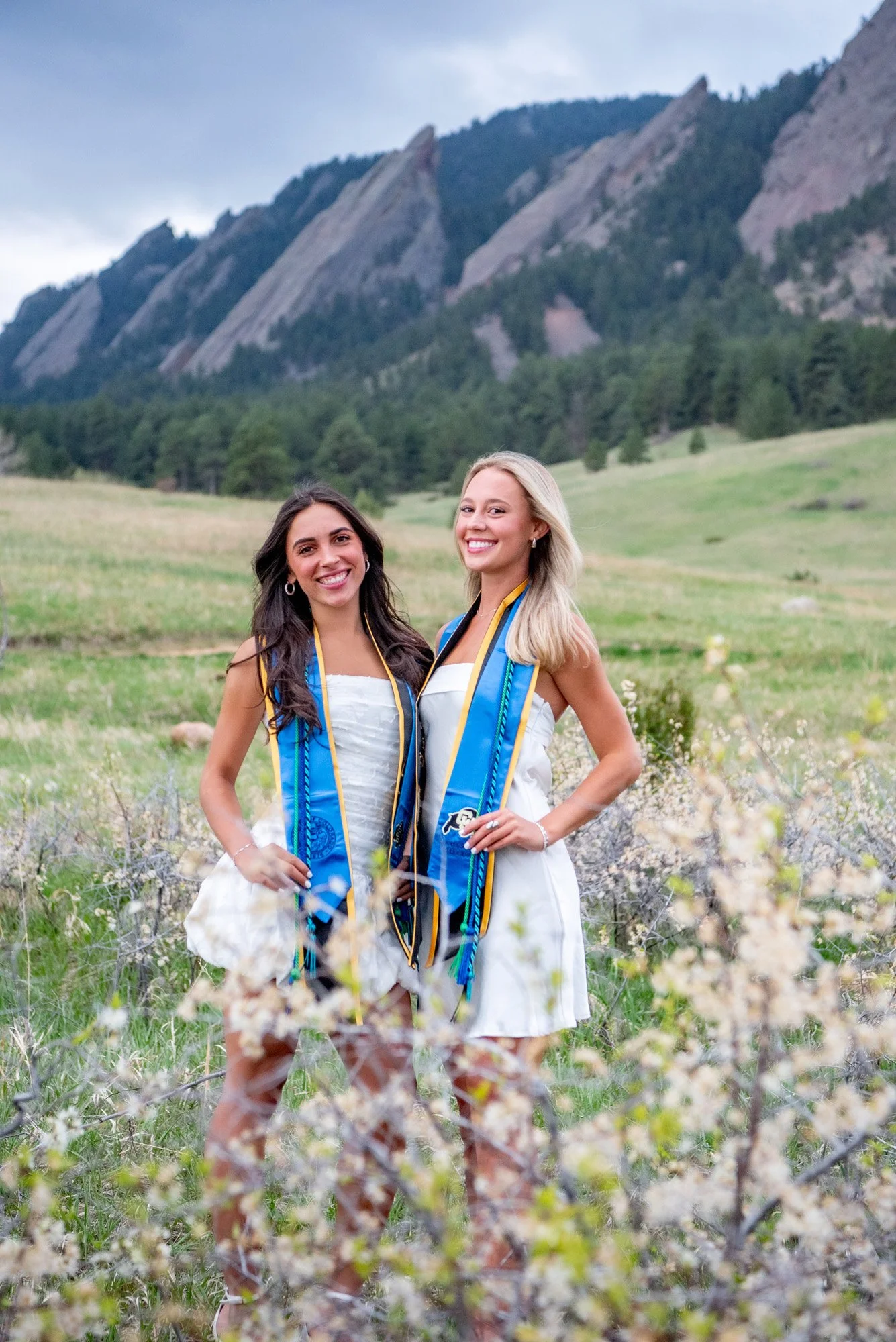Two young women in white dresses celebrating graduation outdoors, wearing blue and gold graduation stoles, with mountains and greenery in the background.