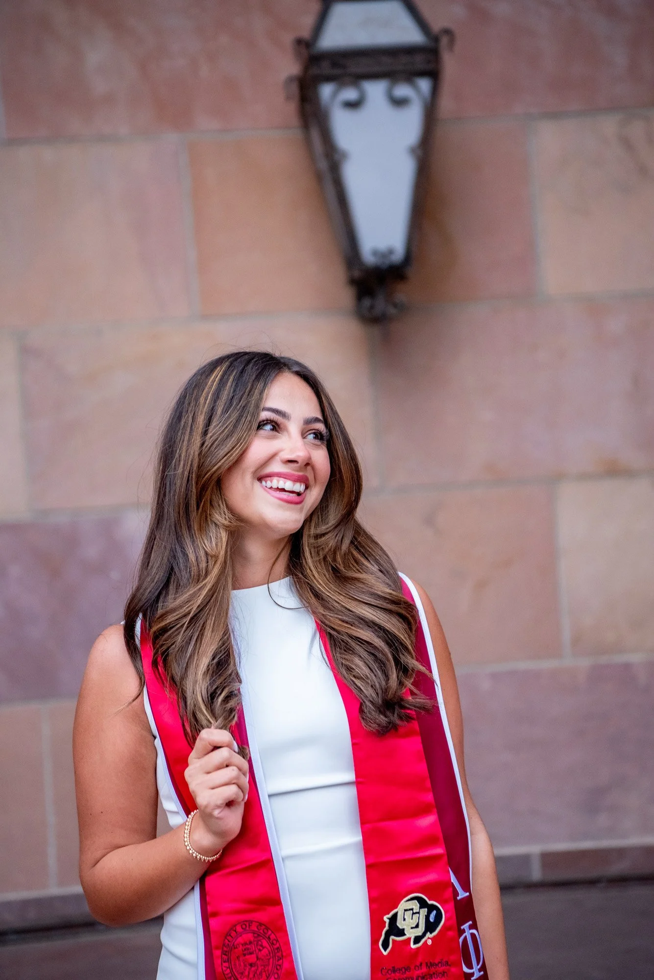 A woman smiling and looking up, wearing a white dress and a red honor stole with logos, standing in front of a stone wall with a vintage black lantern above her.