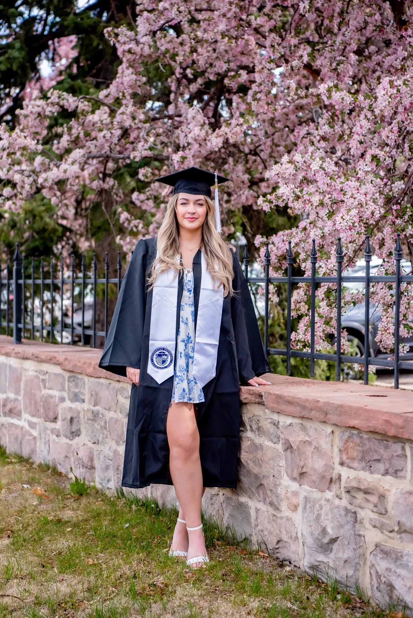 A young woman in a graduation cap and gown standing outdoors next to a stone wall, with pink flowering trees in the background.