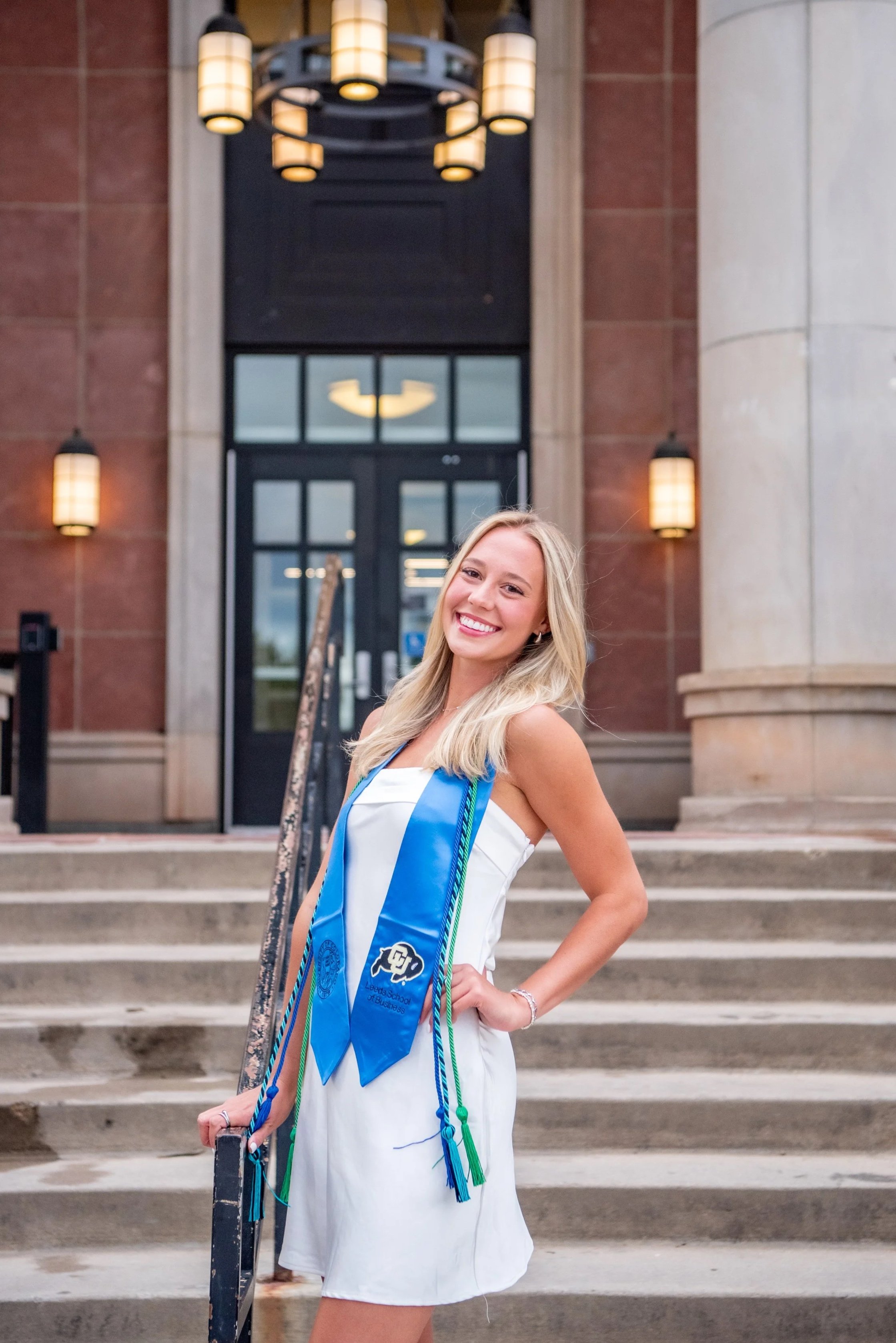 A young woman in a white dress stands on steps in front of a building entrance, wearing graduation cords and smiling.