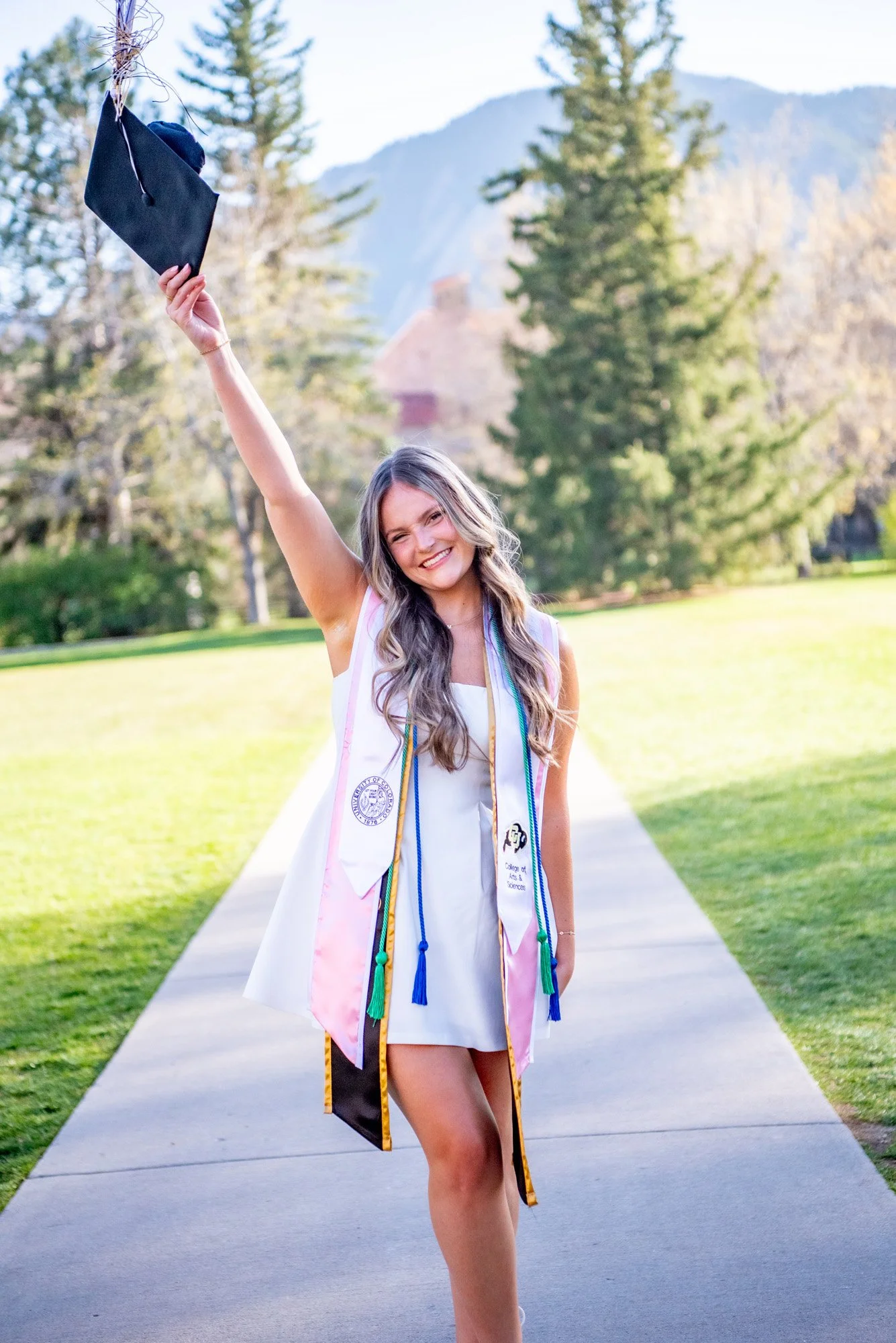 A young woman in a white dress standing on a sidewalk with a graduation cap in her raised hand and wearing graduation stoles and cords, outdoors with trees and mountains in the background.