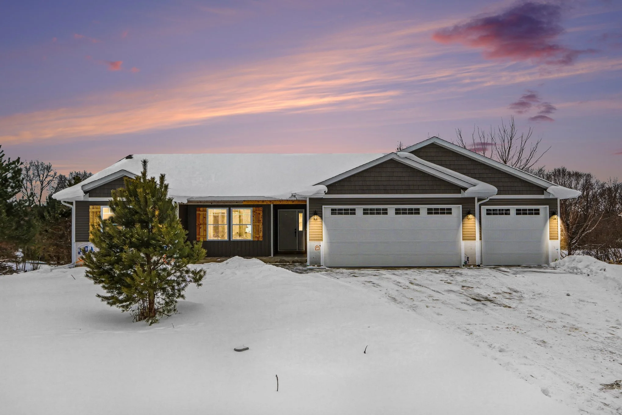 Brand new single-story home by Cornerstone Construction at 8683 Chamomile Drive in Augusta, Michigan, featuring a three-car garage, snow-covered front yard, and warm exterior lighting at sunset, represented by the Jason Veenstra Team | eXp Realty.