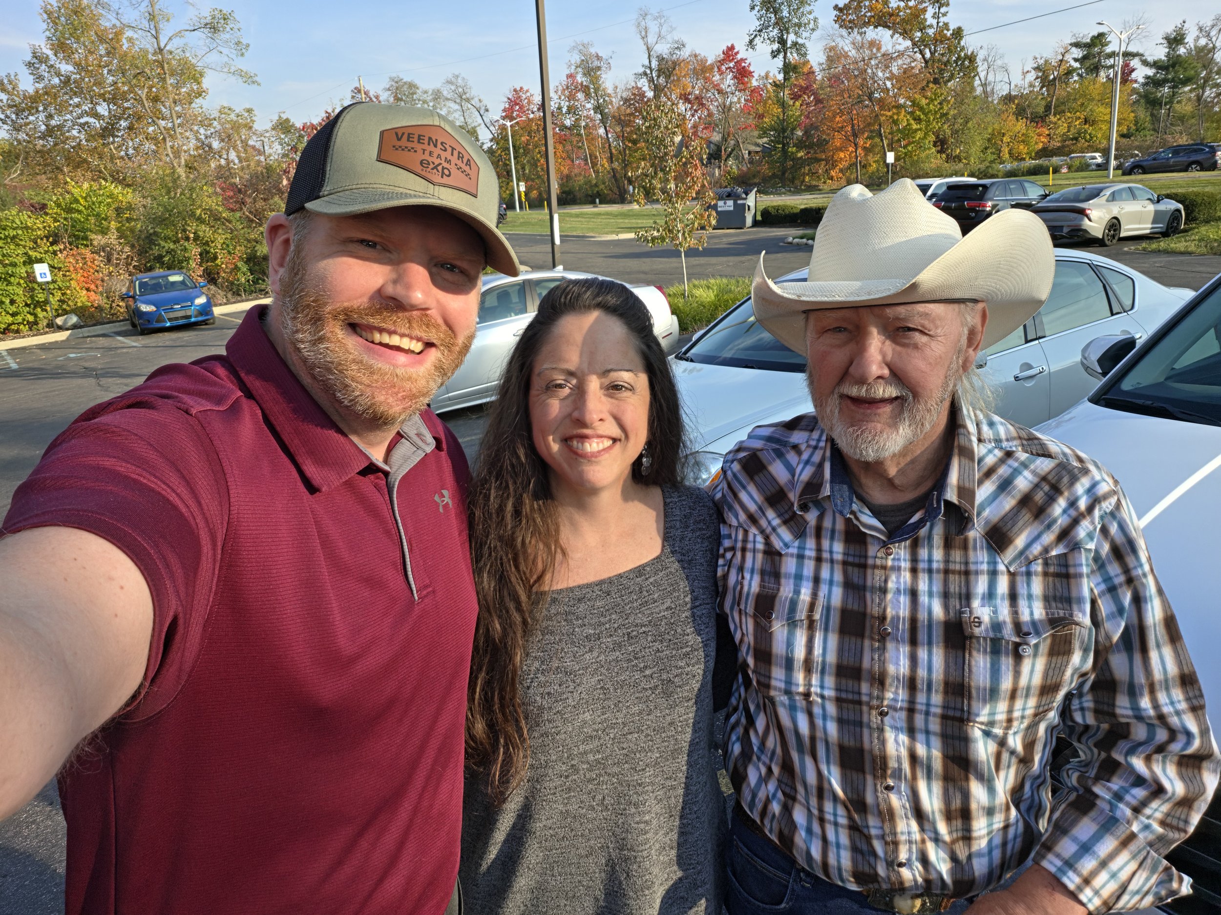 Jason Veenstra, Sarah and Marty smiling outdoors after closing on her parents’ Paw Paw home, with colorful fall trees behind them.