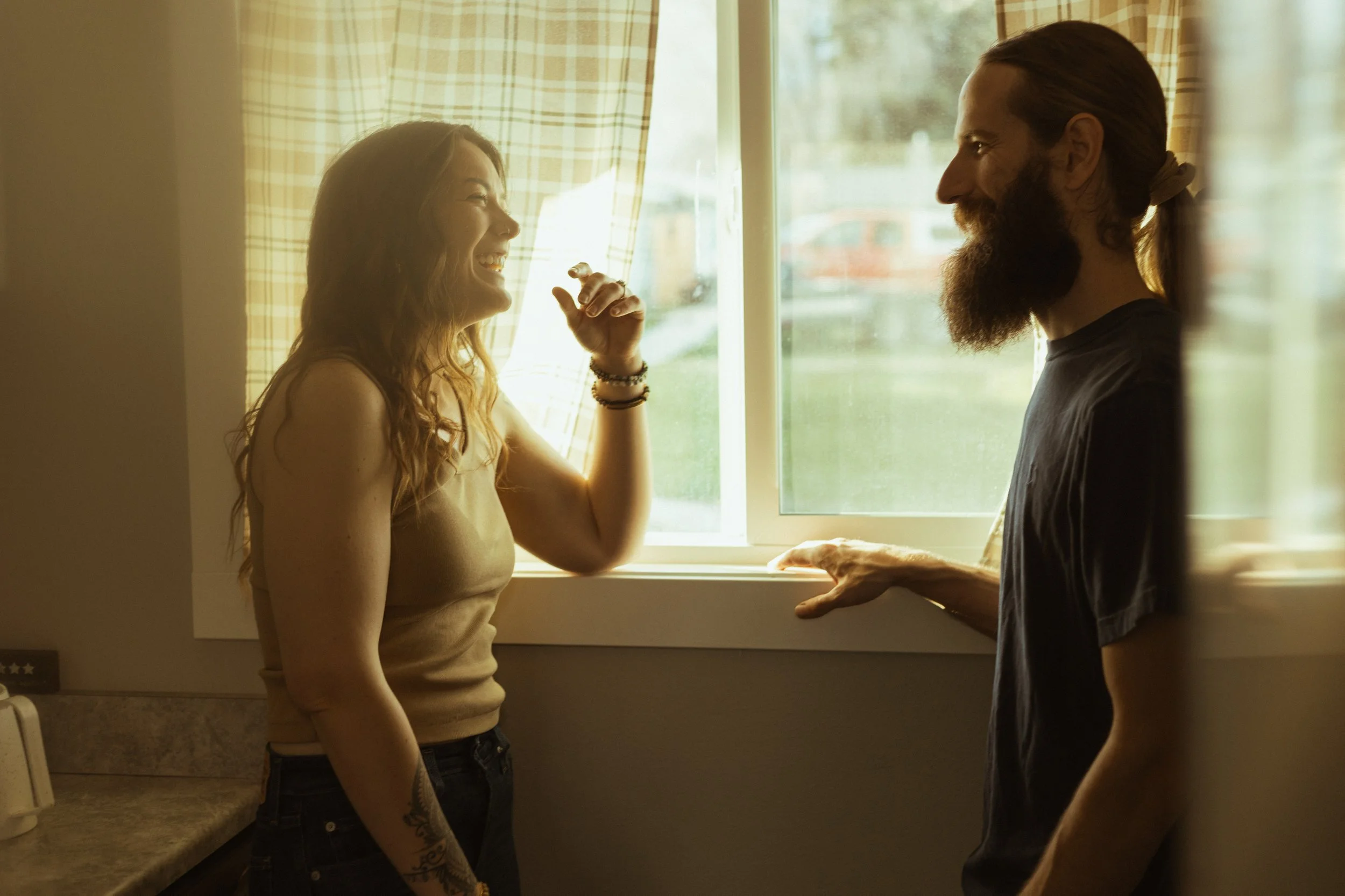 A man and a woman are smiling and talking to each other inside their home near a window with sunlight. The woman has long wavy hair and tattoos on her arm, and the man has a beard, long hair tied back, and is wearing a dark T-shirt.