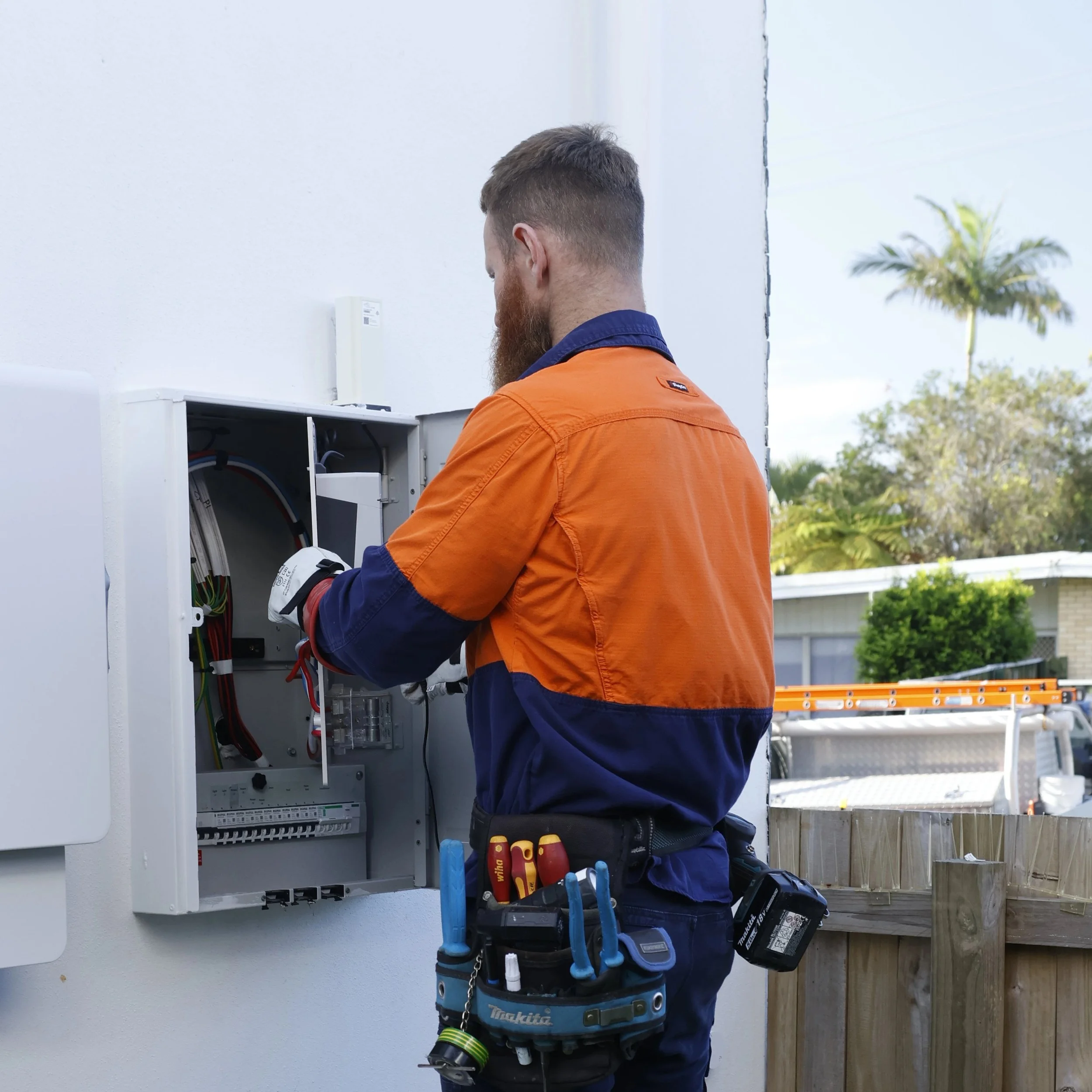 Steven Hord, Master Electrician working on an outdoor electrical panel, wearing tools on belt, with trees and house in background.