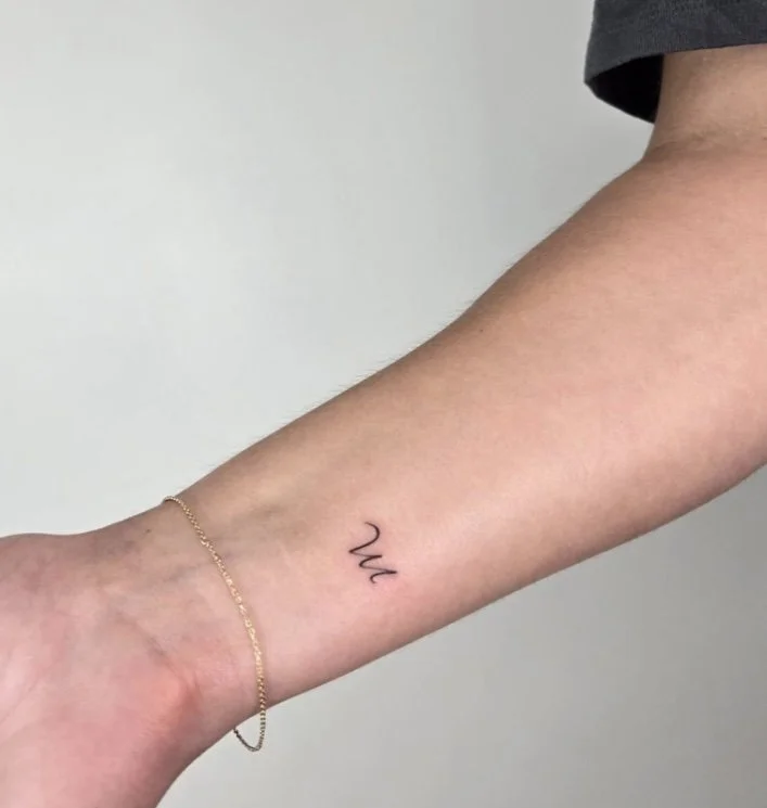 A close-up of a person's forearm with a small, black, cursive tattoo that reads 'Um' and a delicate gold chain bracelet.