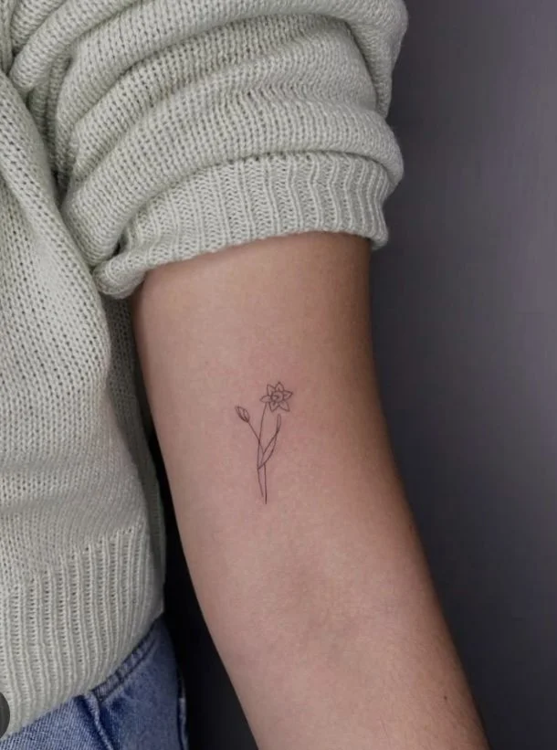 A person's upper arm with a small, minimalist line drawing of a flower tattoo.