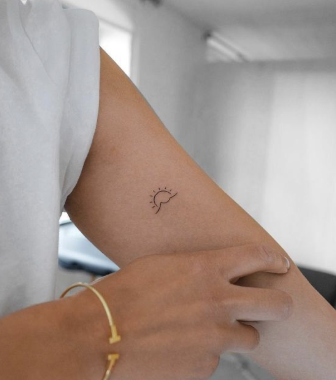 Close-up of a person's arm with a small sun and wave tattoo, with arm resting on another person's hand, in a bright indoor setting.