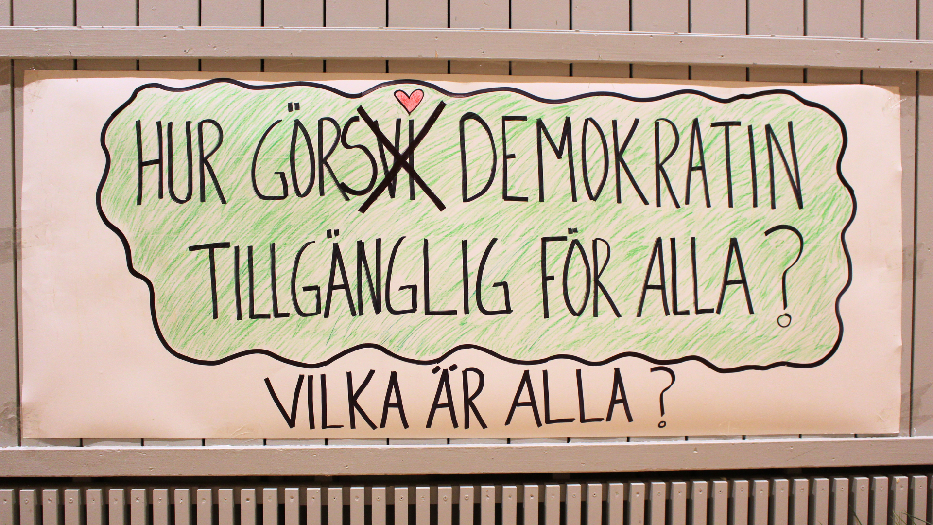 På bild: En hemmagjord skylt med svart text och färger i tusch. Texten säger: “Hur görs demokratin tillgänglig för alla? Vilka är alla?”