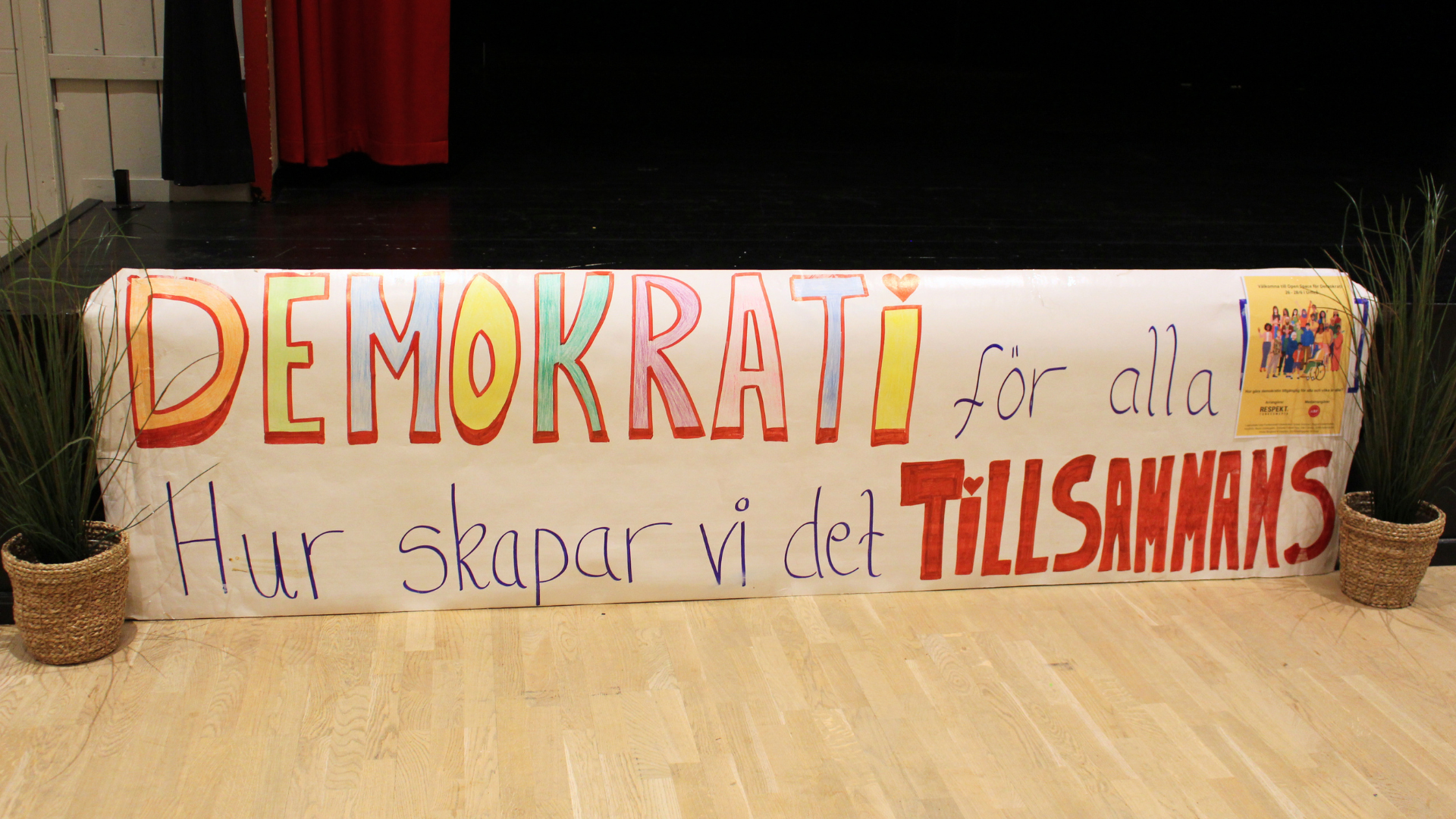På bild: En till hemmagjord skylt i tusch på papper med massa olika fina färger. Texten säger: “Demokrati för alla. Hur skapar vi det tillsammans”.
