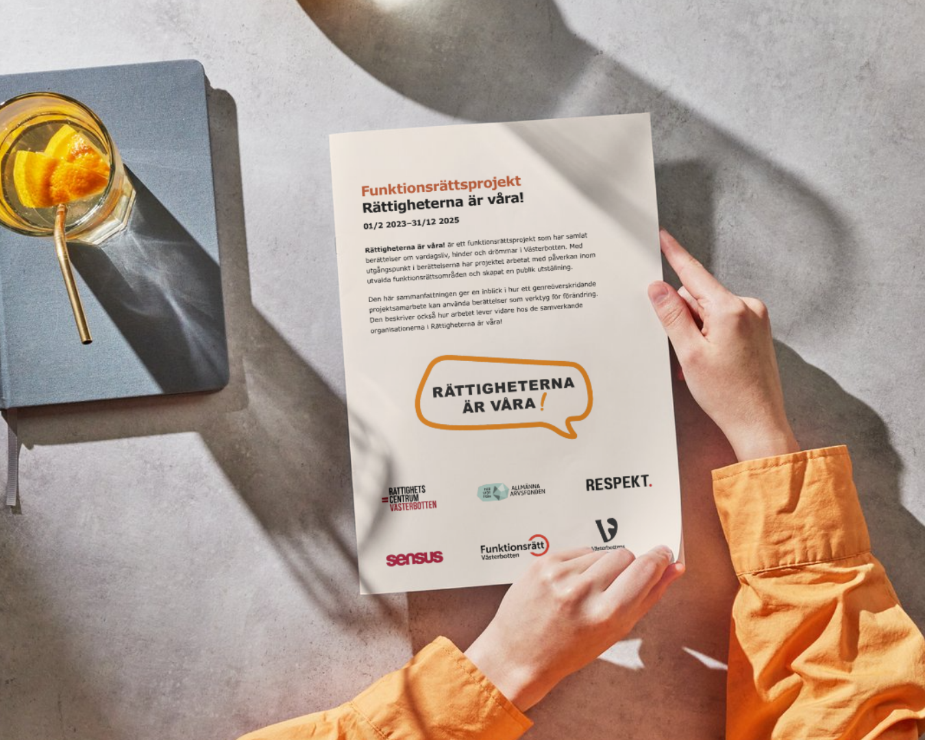 På bild: Två händer med orangea ärmslut håller i slutrapporten över ett bord och håller på att vända blad. Man ser slutrapportens framsida. Till vänster står ett dryckesglas med vatten, sugrör och två apelsinklyftor i.