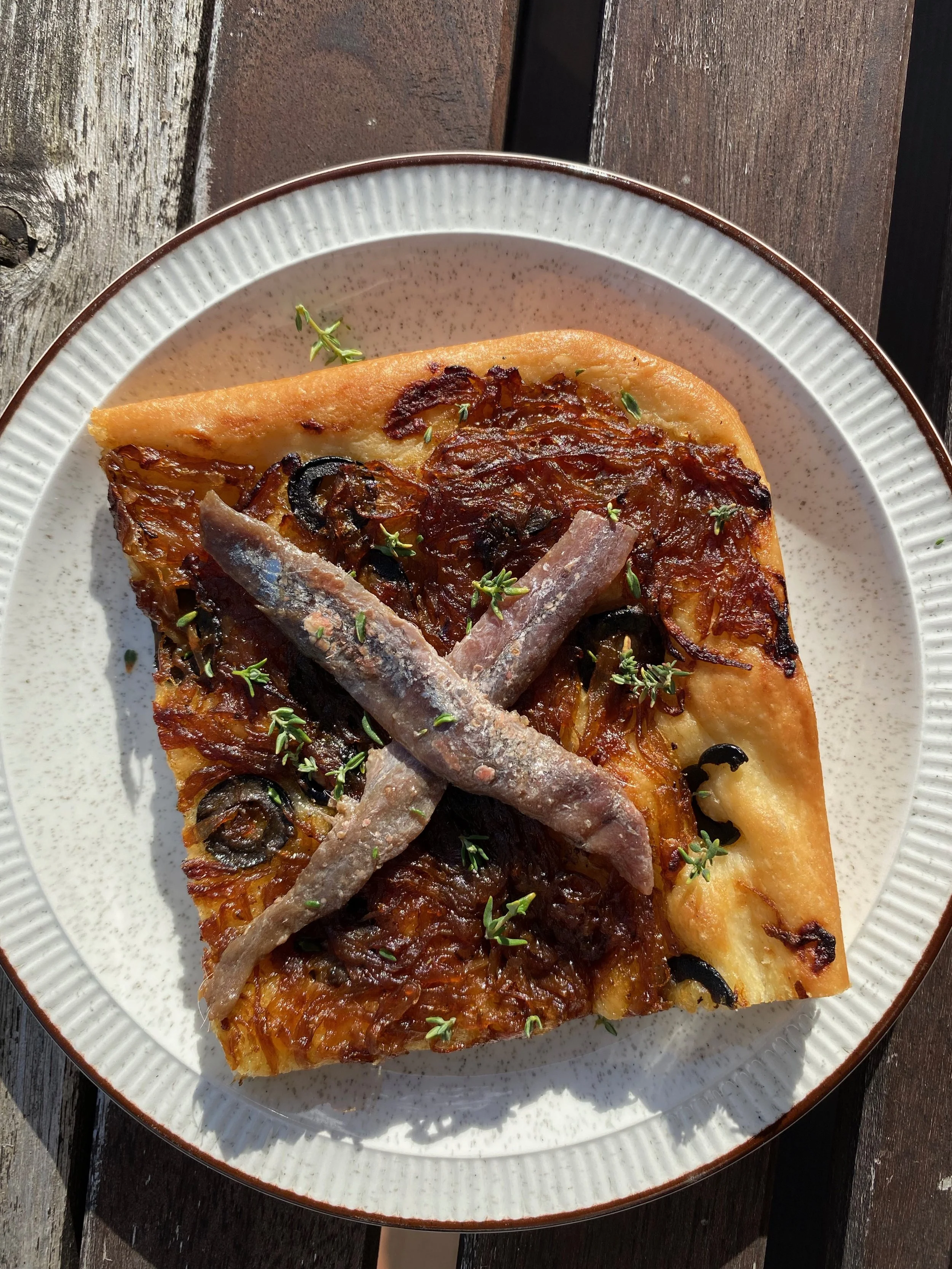 speckled plate with brown trim and goden slice of dough toppped with olives, caramelised onion, anchovies and fresh thyme.