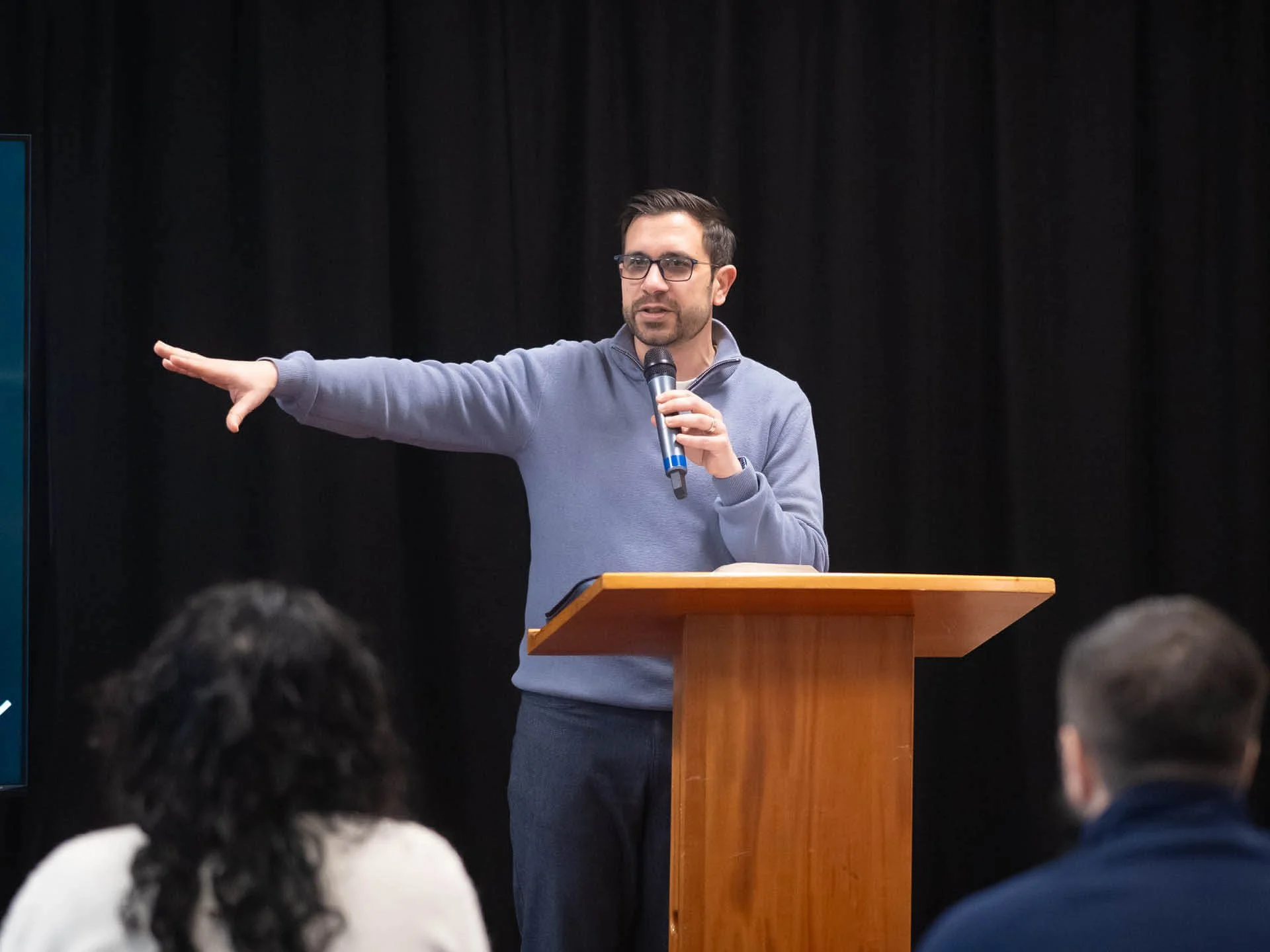 A speaker in a blue jumper stands behind a wooden lectern, holding a microphone with one arm outstretched.