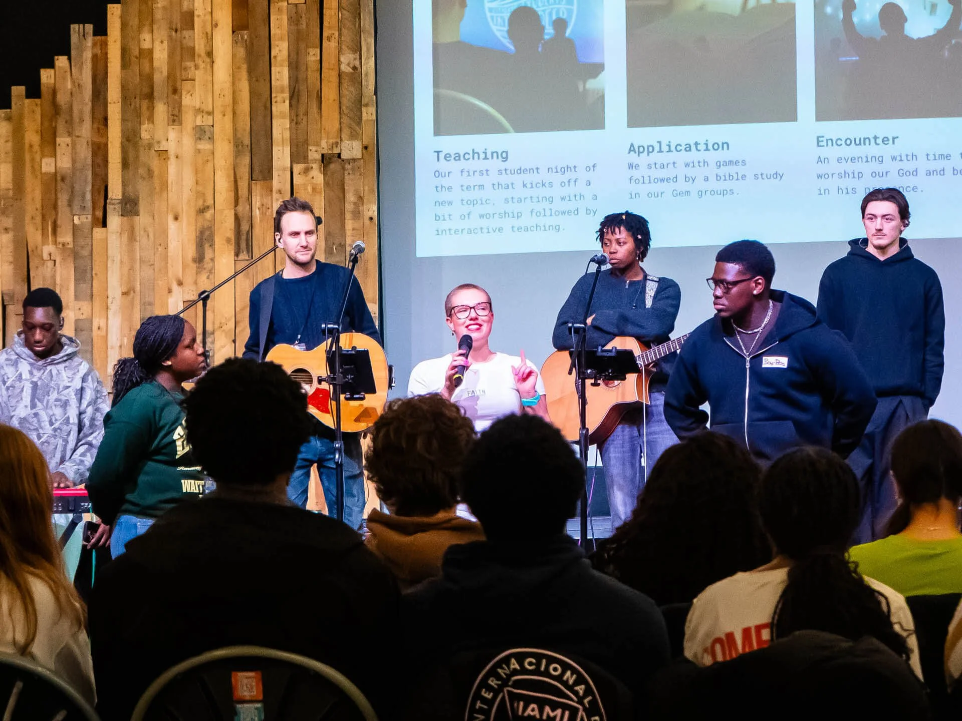 A young female student speaks enthusiastically to a crowd of young people, with a student-led band on stage behind her.