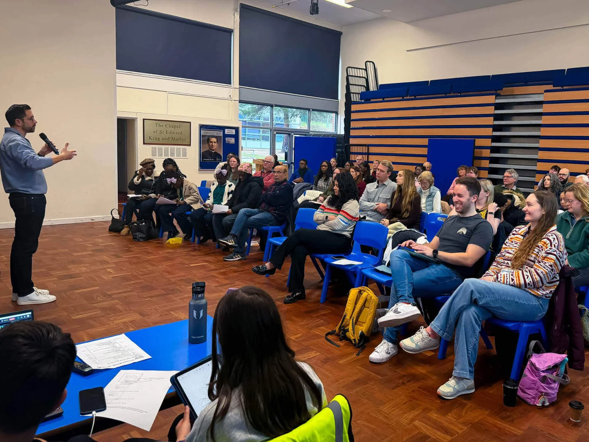 A diverse, smiling congregation gathers in a school hall, listening to a man in a blue jumper holding a microphone.