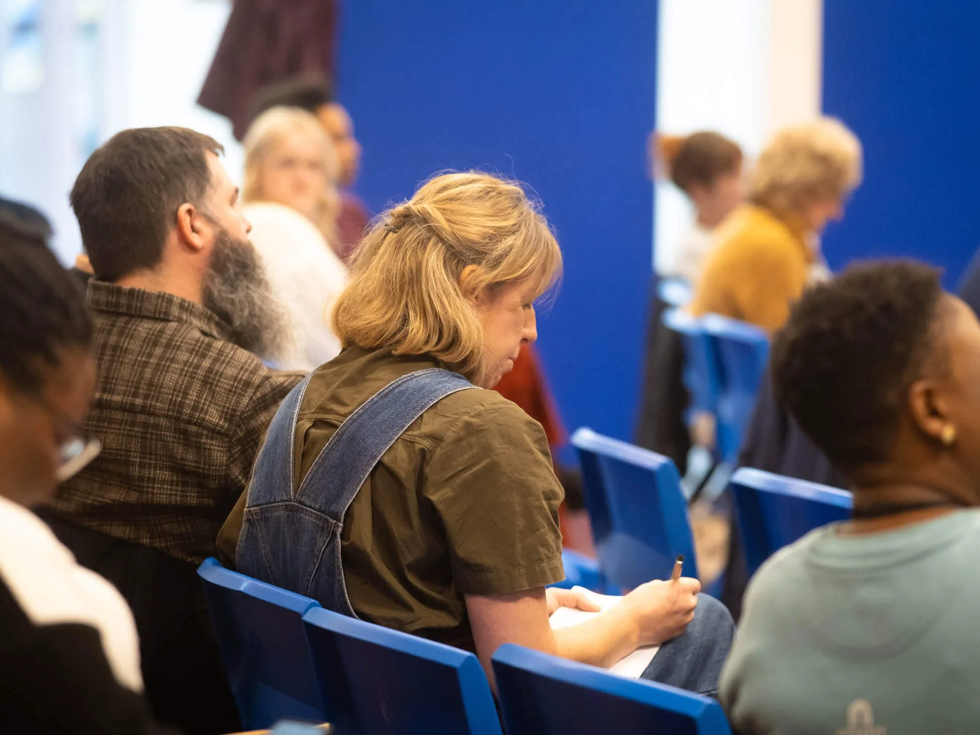 A woman in dungarees sits in a church congregation next to her husband, writing on a notepad.