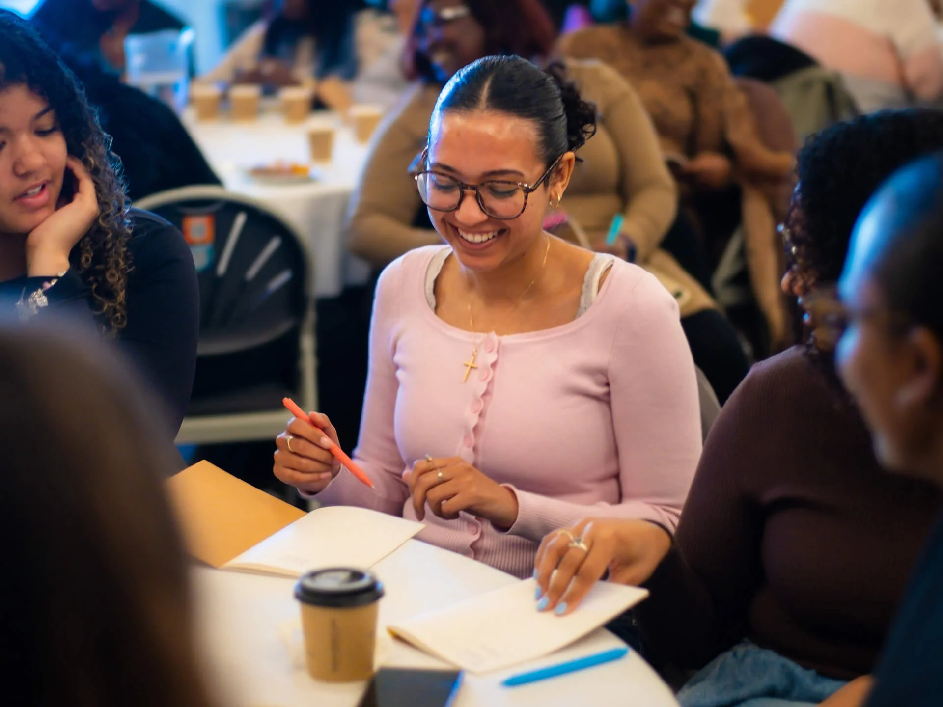 A multicultural group of female students and young adults sitting around a table discussing the Bible in community, while the girl at the centre of the photo smiles and holds a pen, preparing to write her thoughts and reflections in a notebook.
