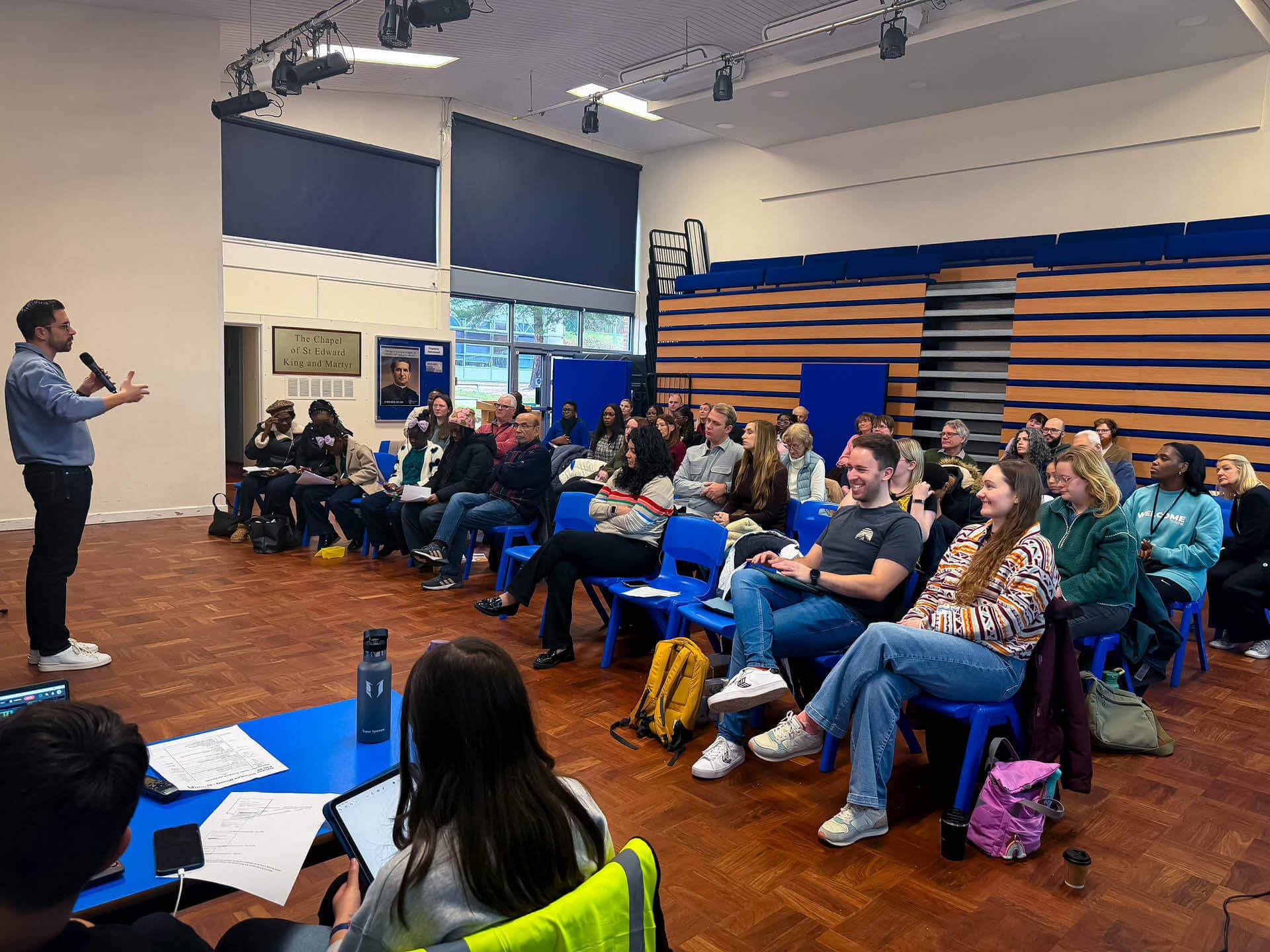 A diverse, smiling congregation gathers in a school hall, listening to a man in a blue jumper holding a microphone.