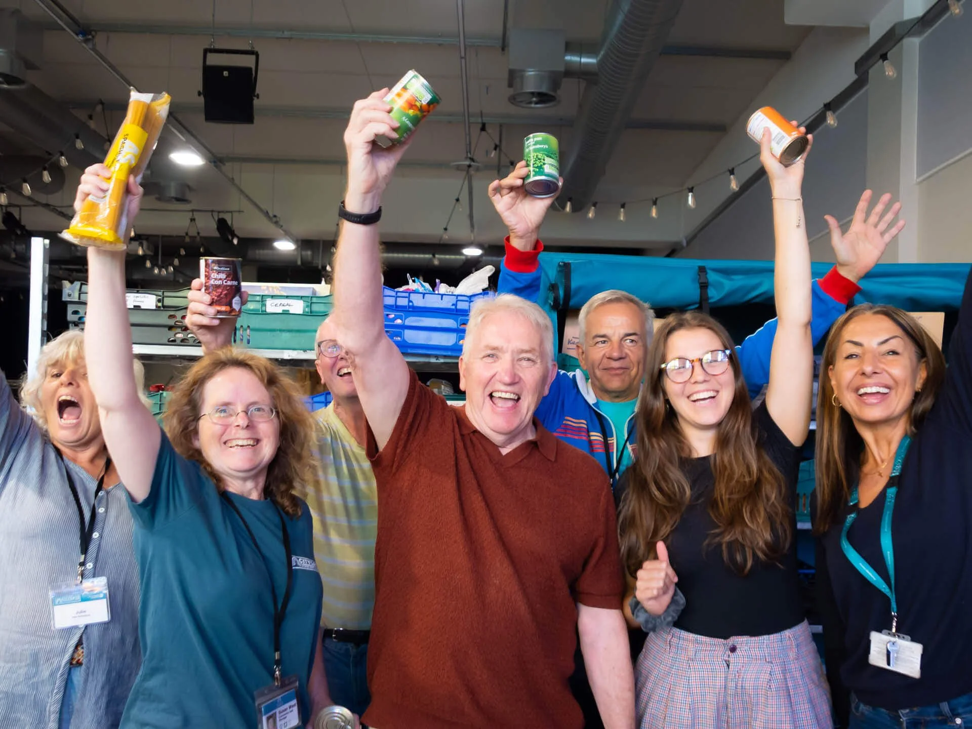 A cheerful group of community hub volunteers stand in front of stacked crates of food, smiling as they hold up tins of fruit and vegetables.