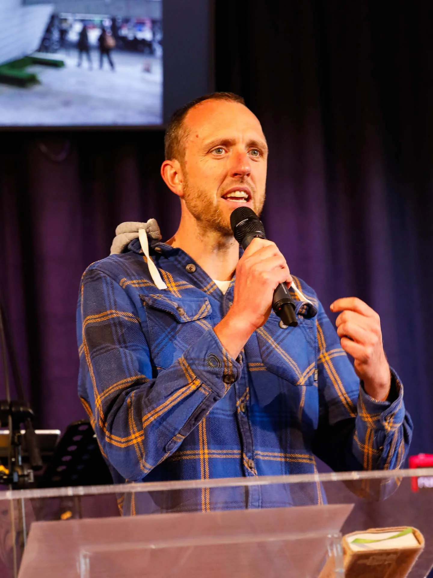 A man in a blue checkered shirt stands confidently behind a lectern, speaking into a microphone.