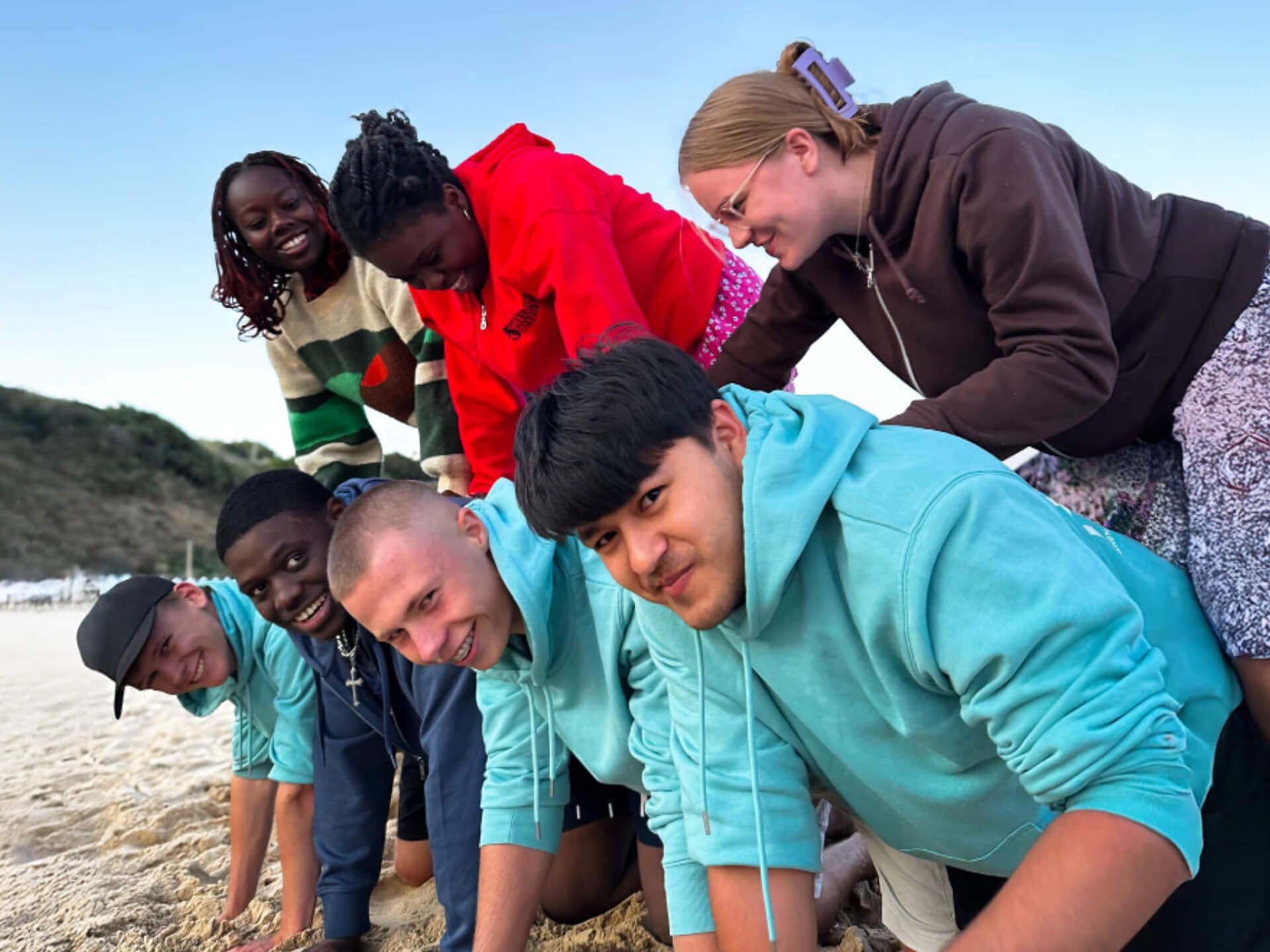 A multicultural group of students and young adults forming a human pyramid on Bournemouth beach, climbing over one another while laughing and smiling, symbolizing community and friendship.