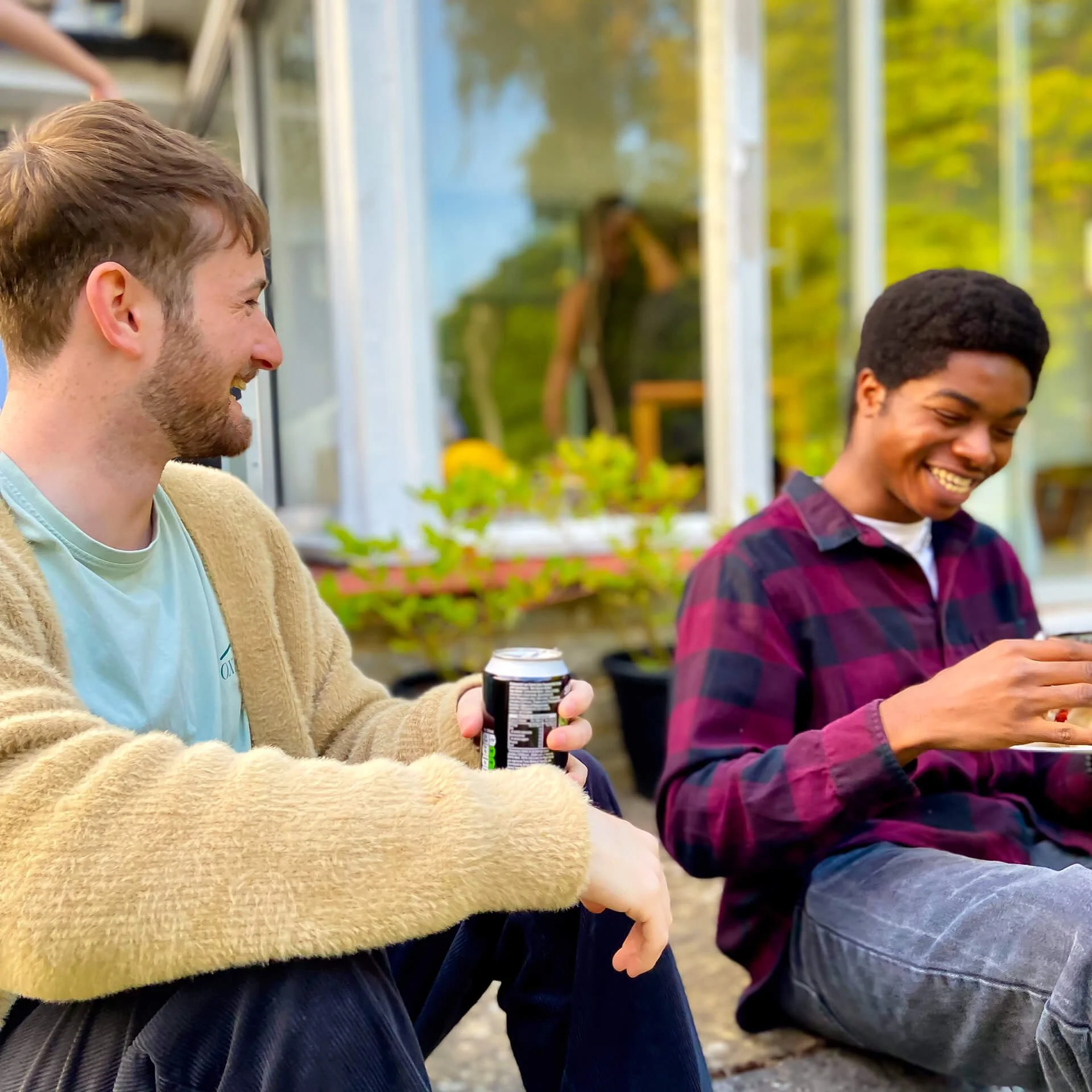 Two young men at a barbecue, sat next to each other, laughing , chatting, and connecting with one another.