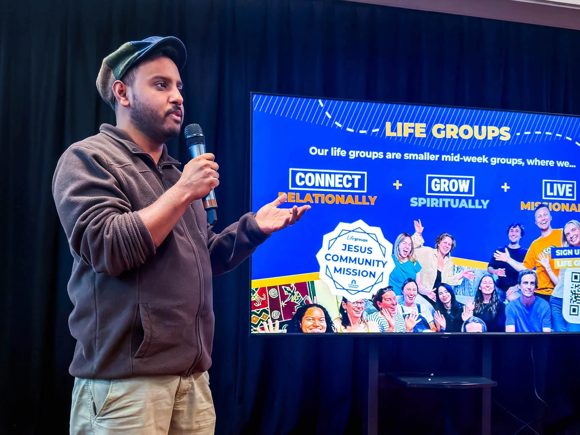 Young man wearing a cap holds a microphone at the front of a room, speaking animatedly about church life groups.
