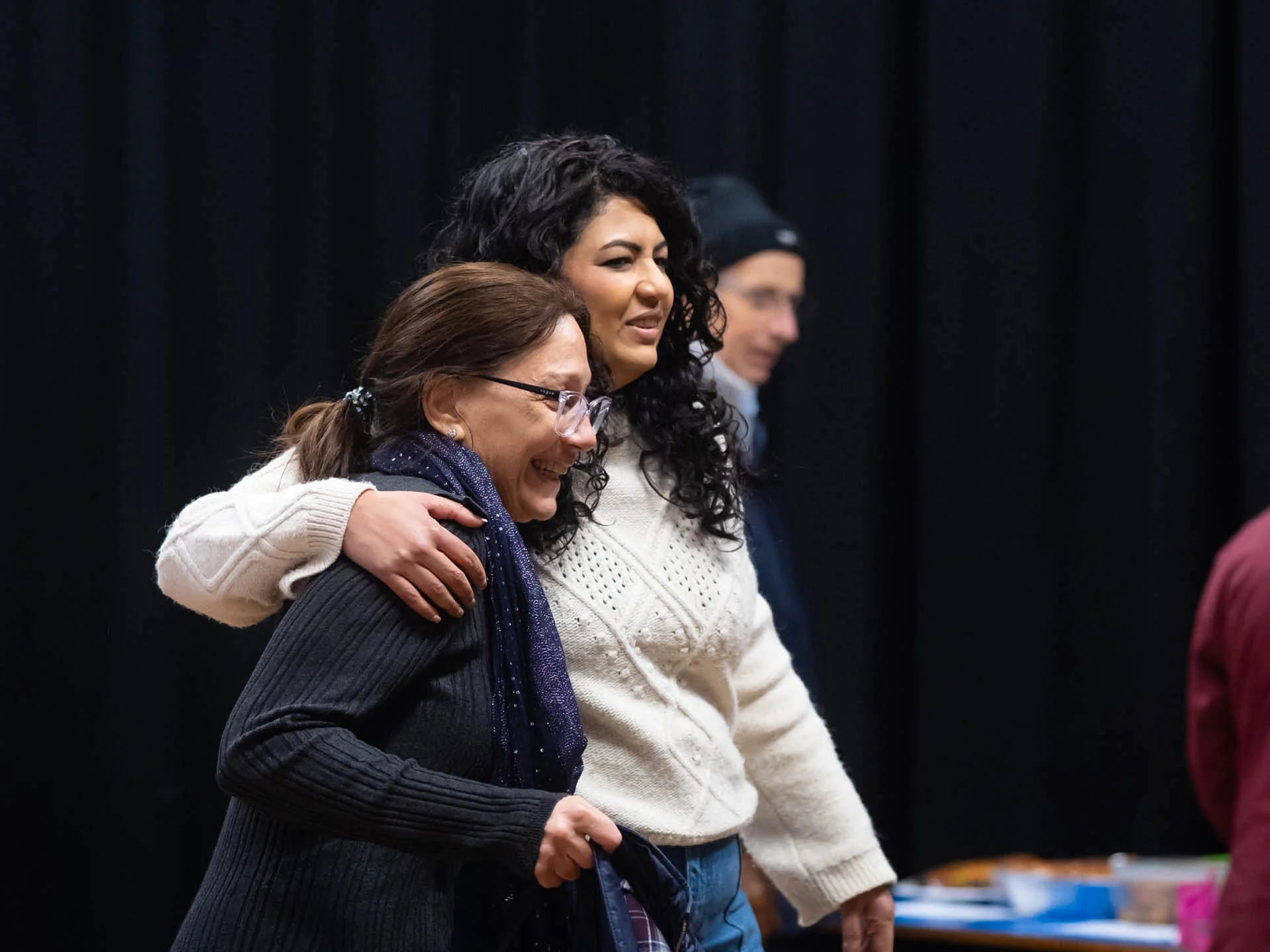 A woman in a white jumper stands with her arm around another smiling woman.
