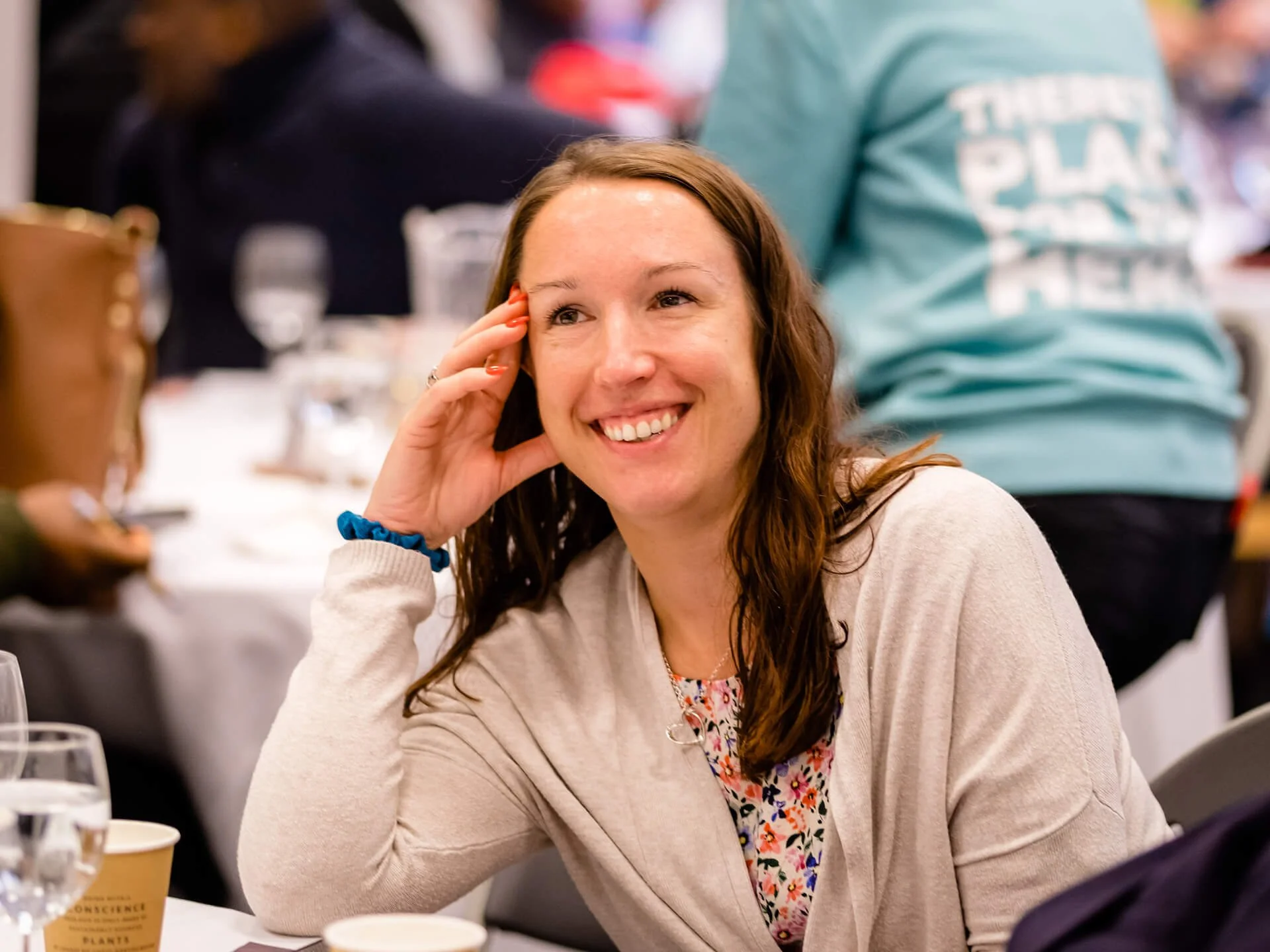 A smiling middle-aged lady is sat at church around a table, listening to someone speaking and learning.