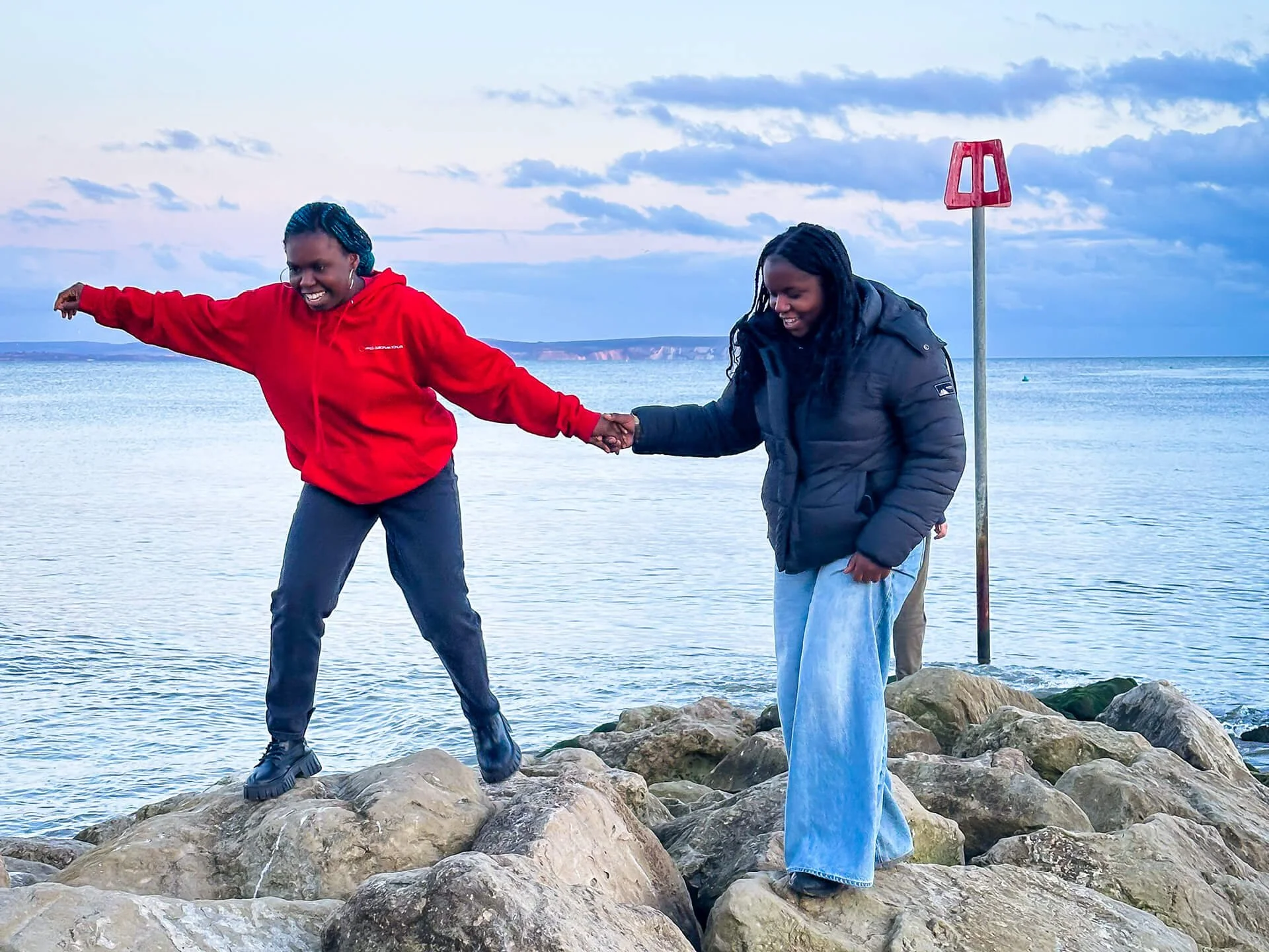 Two young women at the beach, smiling, holding hands and helping one another walk over some rocks.