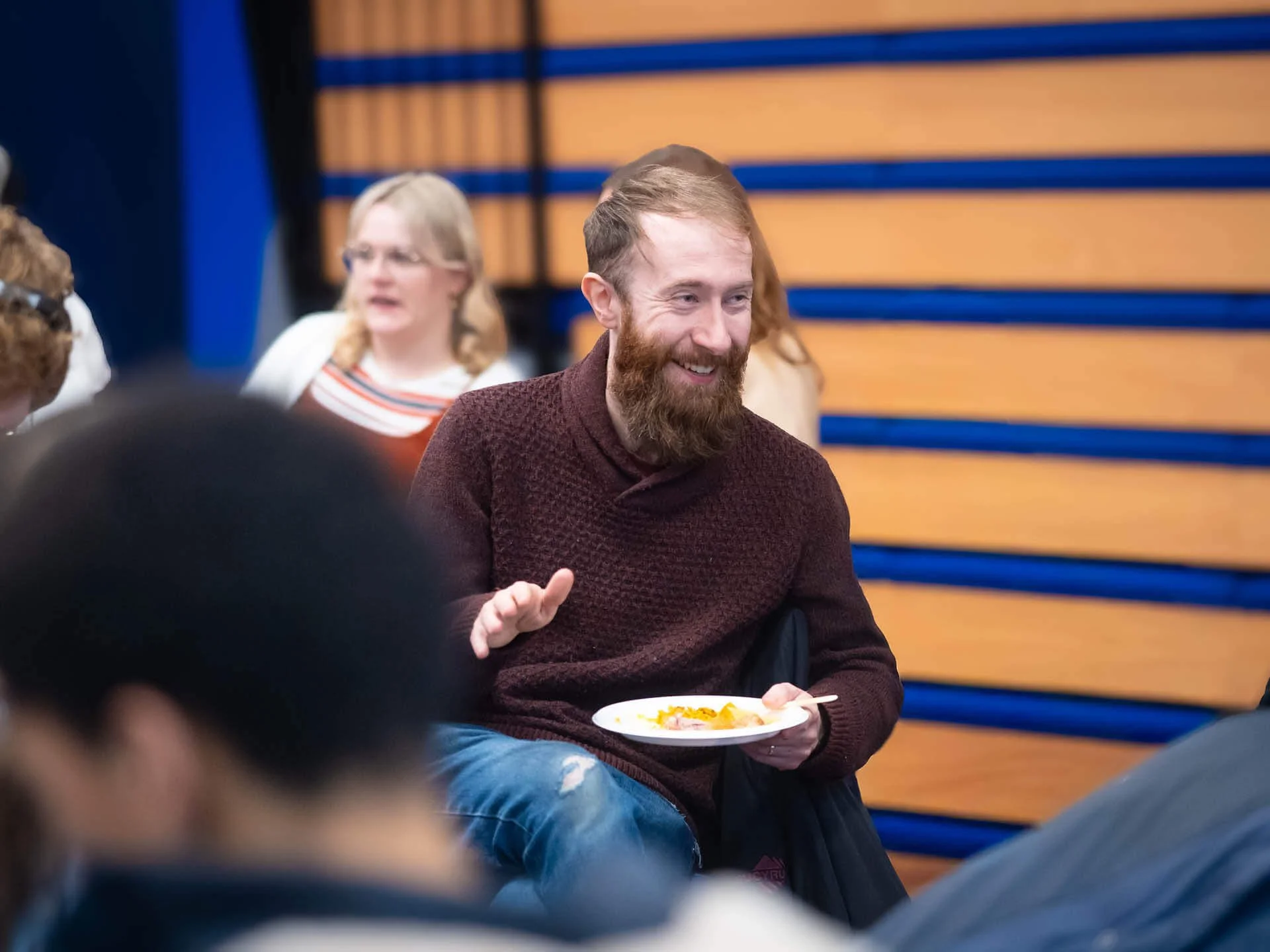 A man wearing a maroon jumper sits holding a plate of food, smiling animatedly while engaged in conversation with someone.”