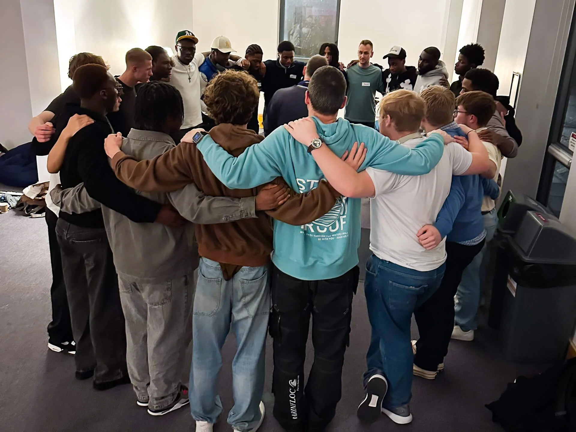 A large, multicultural group of male Christian students and young adults in their twenties standing in a circle with their arms around each other’s backs, as a man in the middle teaches and declares truth from the Bible over them.