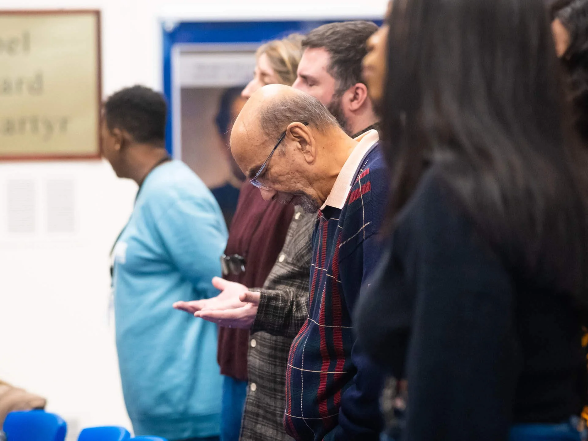 An elderly man wearing a plaid jumper stands in a church congregation with his head bowed in reverence.