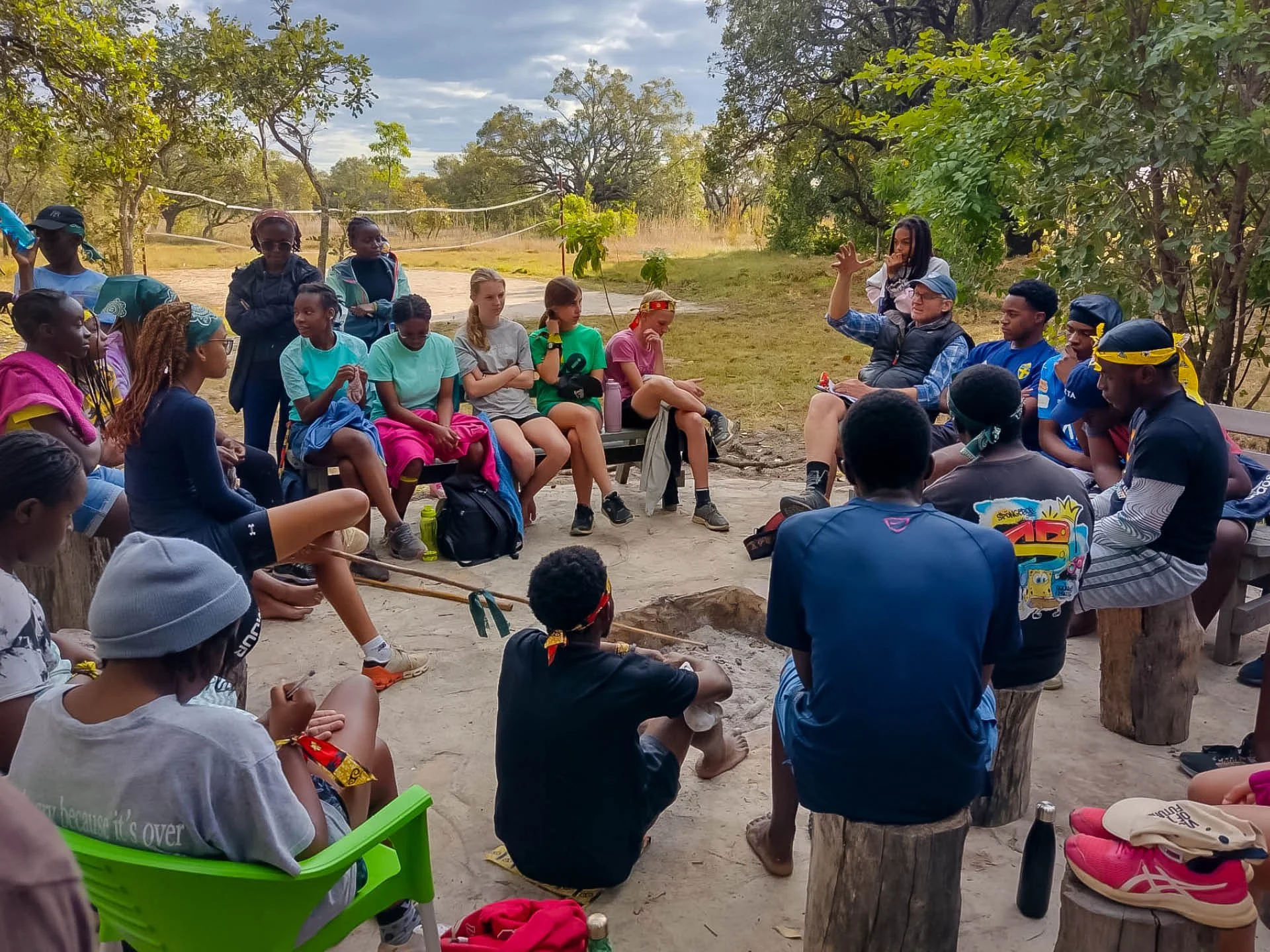 A diverse group of young people in Zambia sit gathered around a campfire, listening attentively as an animated speaker addresses them.