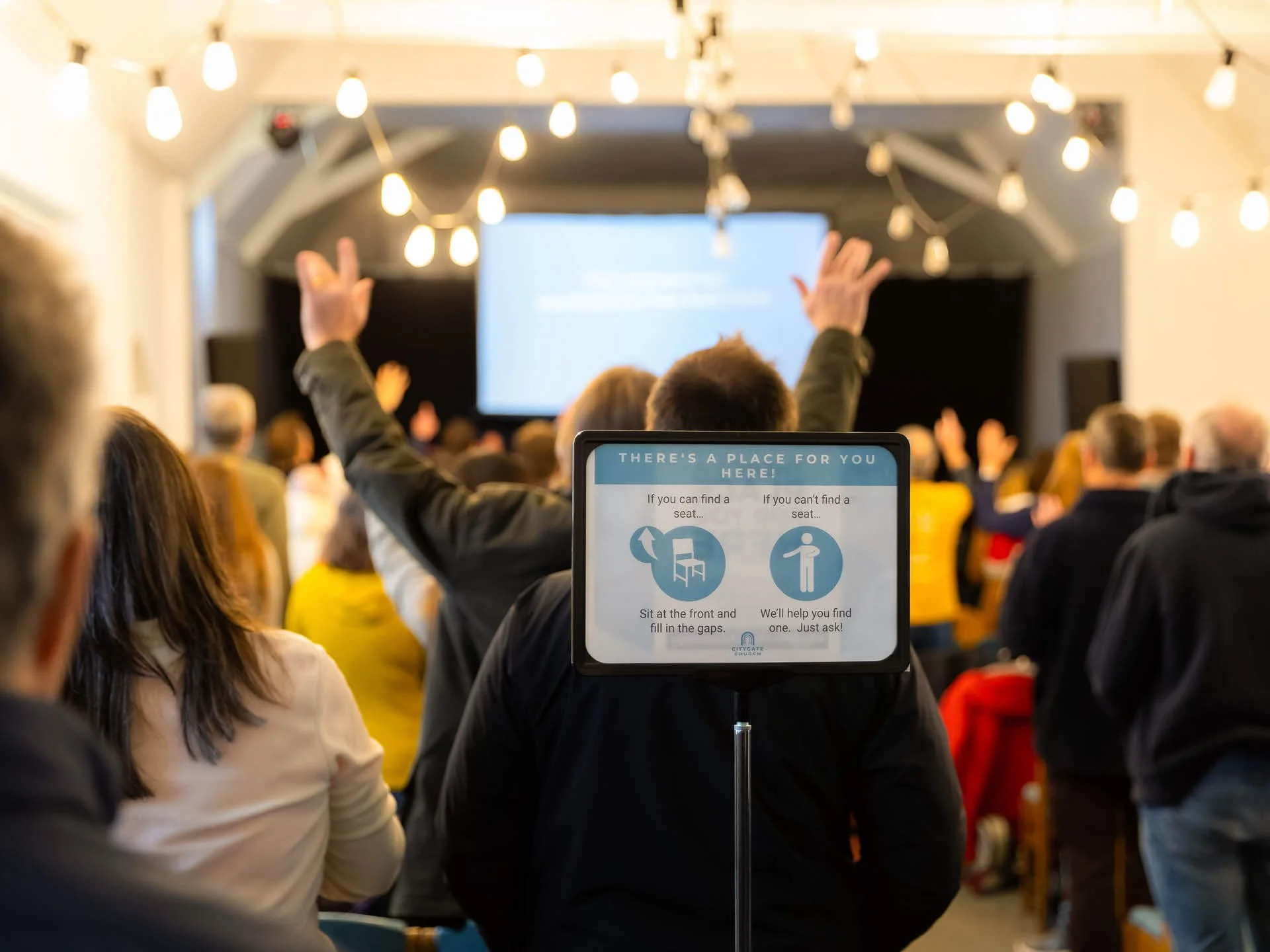 A lively congregation worships with their hands raised. The ceiling is decorated with hanging lights, and a sign in the foreground reads, 'There's a place for you here.'
