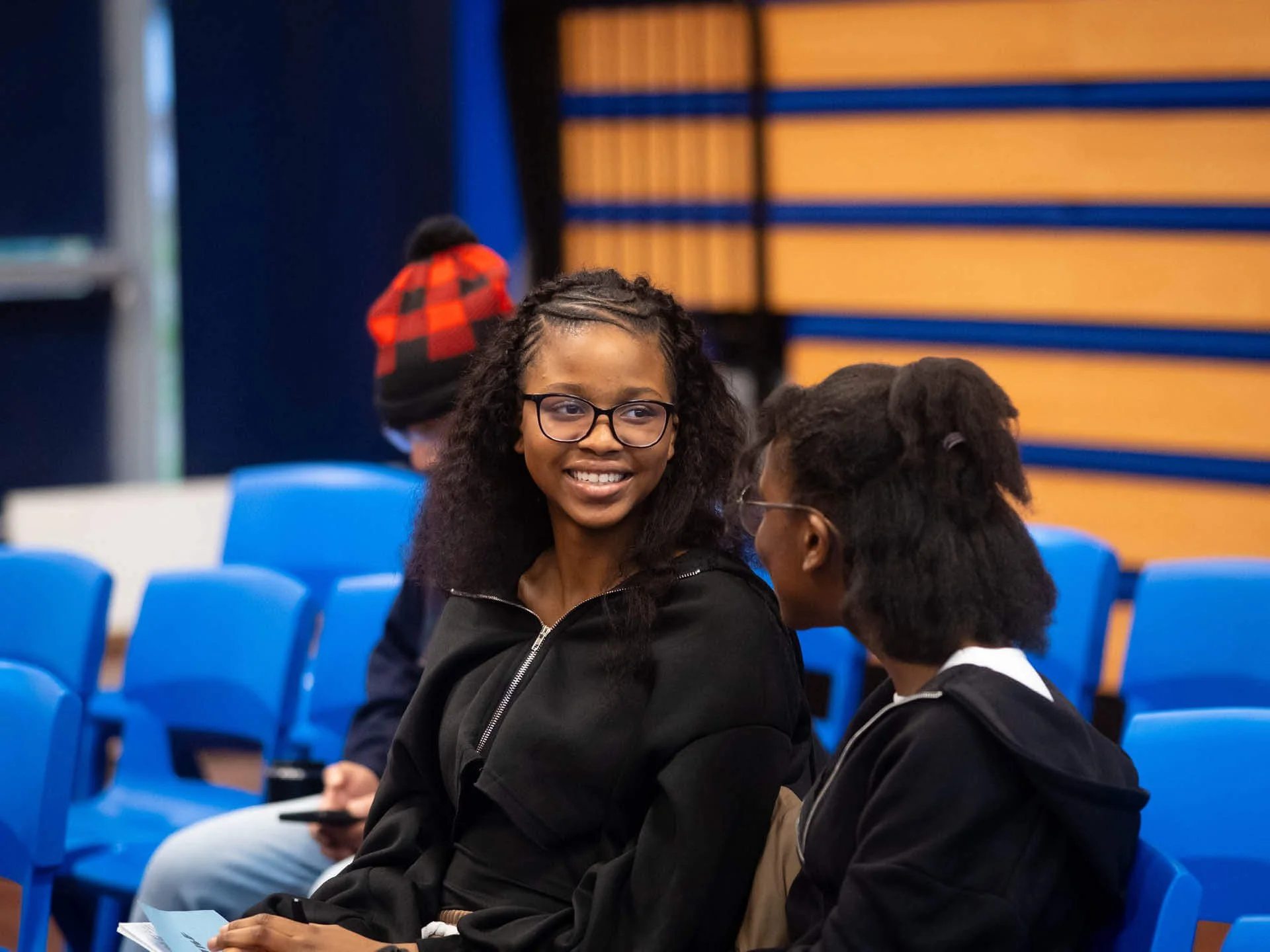 Two teenage girls in black sit in a school hall, smiling and chatting together.