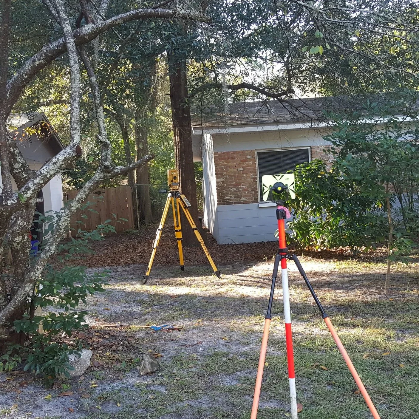 Land survey equipment set up beside a residential structure, with a total station and tripod positioned to measure property boundaries among trees and landscaping.