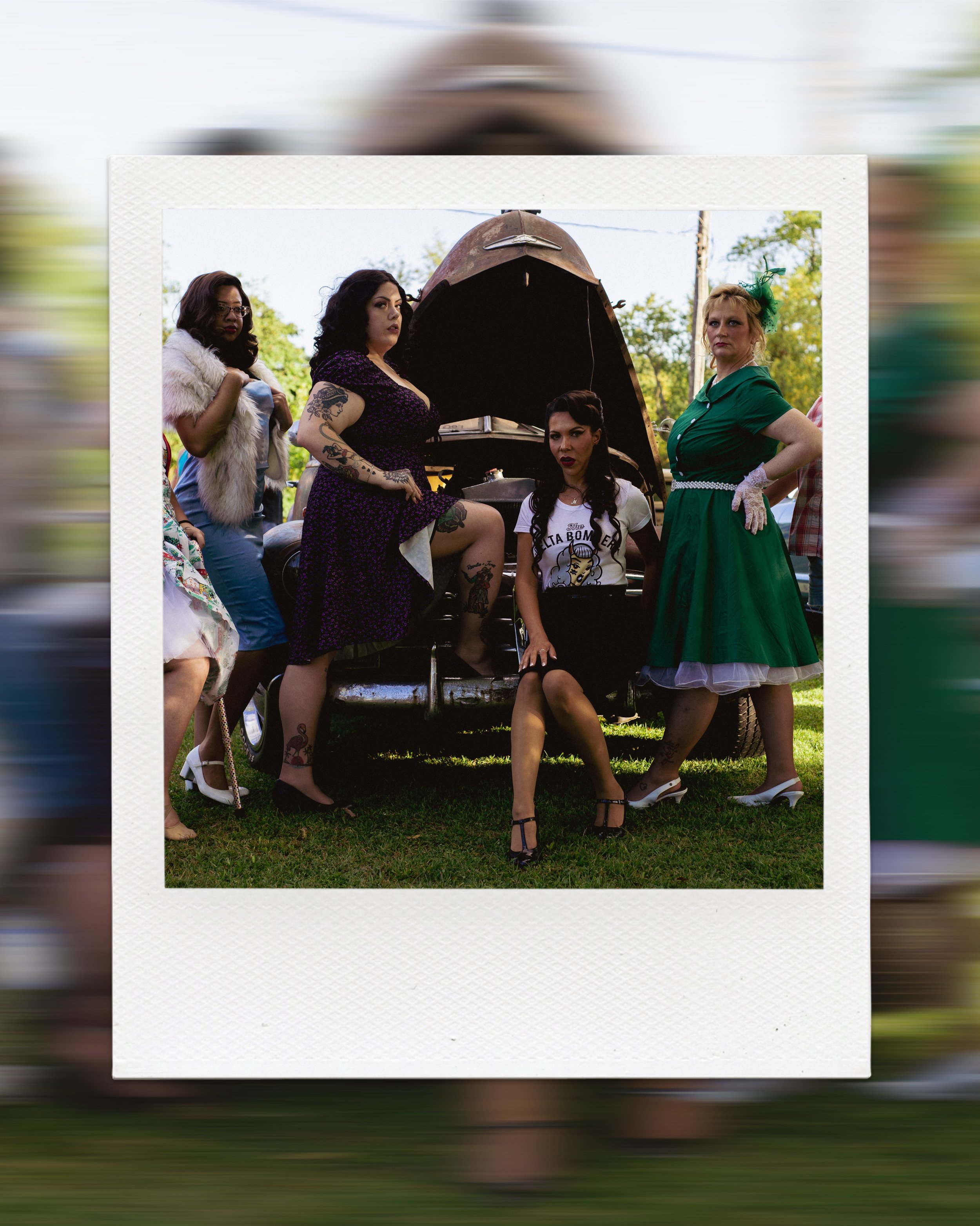 Four women in vintage clothing stand by a classic car with its hood open.