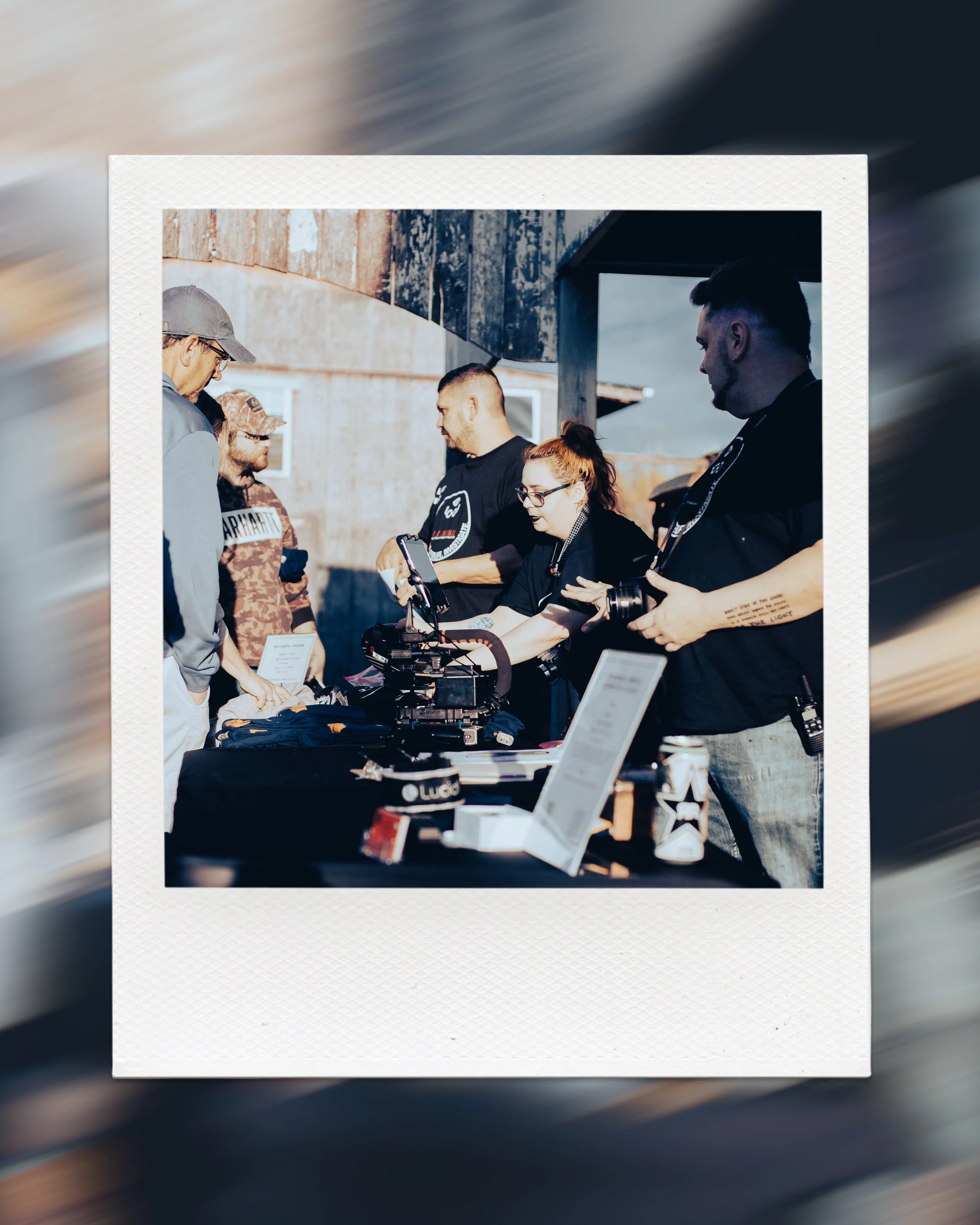 Group of people gathered around a table with camera equipment outdoors, wearing casual clothing, engaging in conversation.