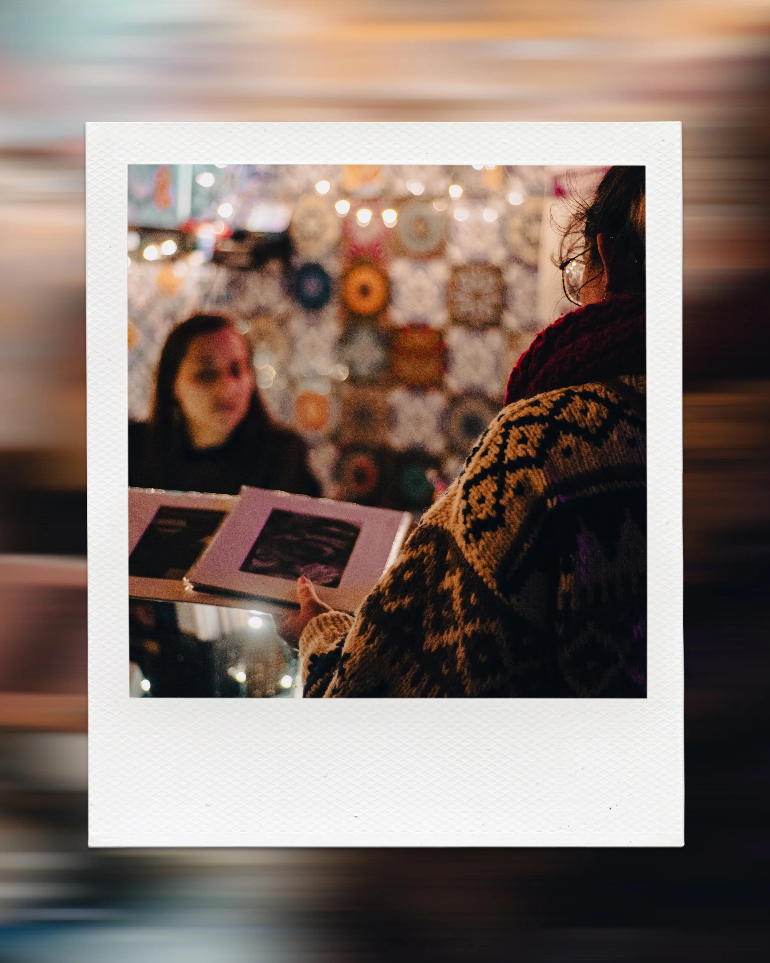 Person in patterned sweater holding art prints at a craft market with colorful background and lights.