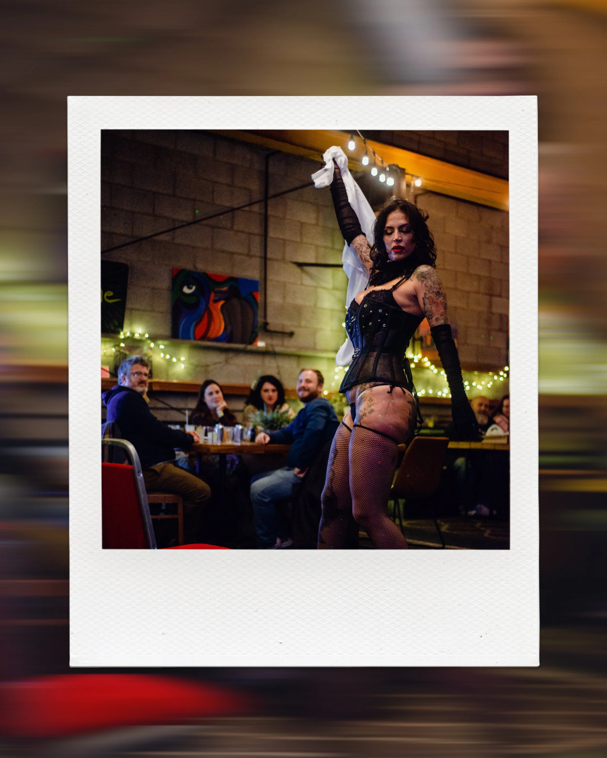 Tattooed performer in black corset at cabaret show with audience seated at tables and art on walls.
