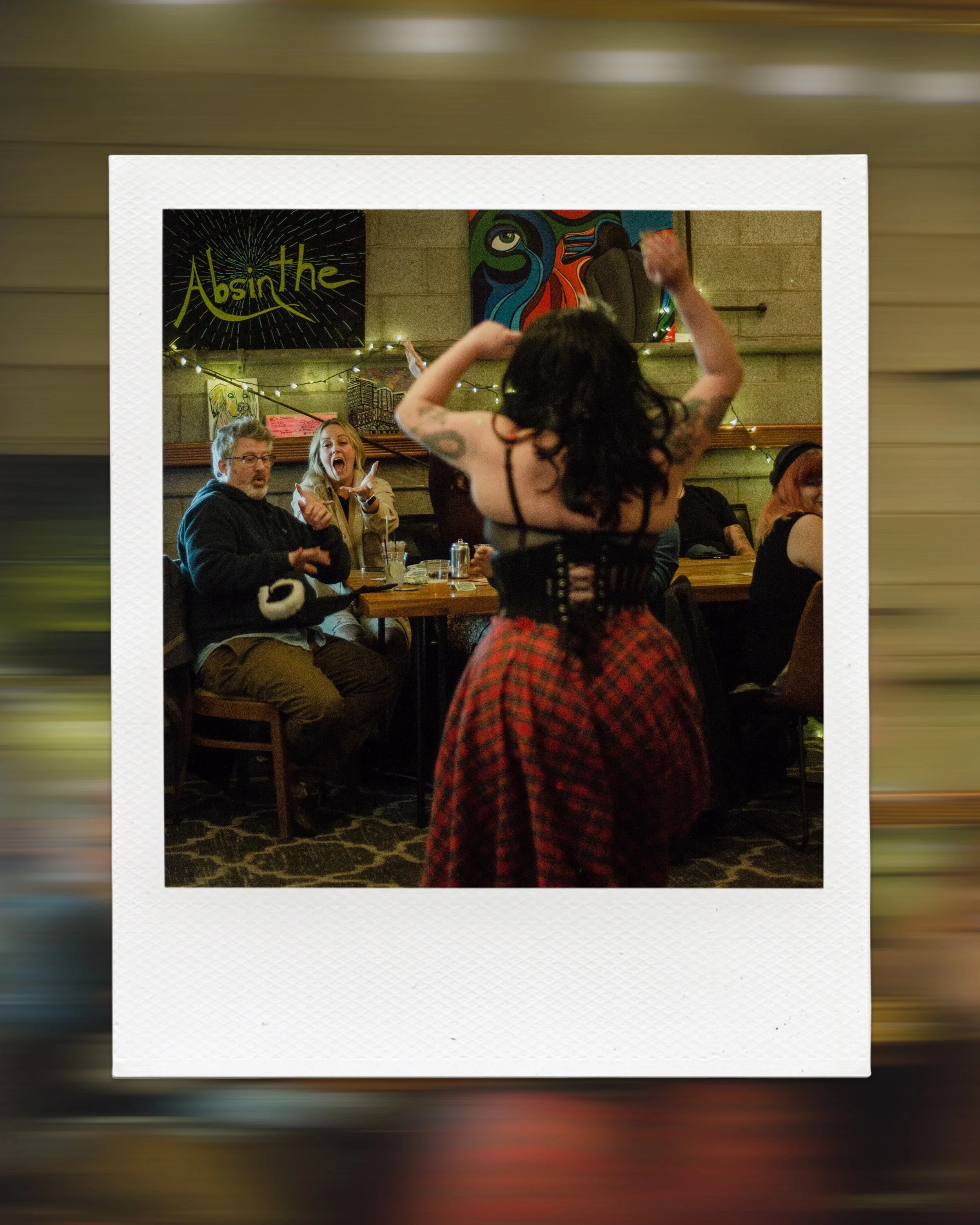 A performer in a red plaid skirt entertains a seated audience at a lively venue, with artwork and a sign reading 'Absinthe' in the background.