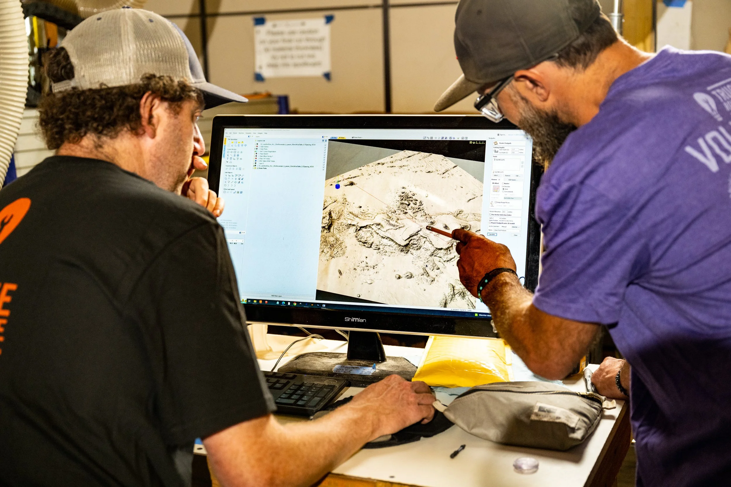 Two men analyze a 3D model or terrain map on a computer screen, with one pointing at the model.