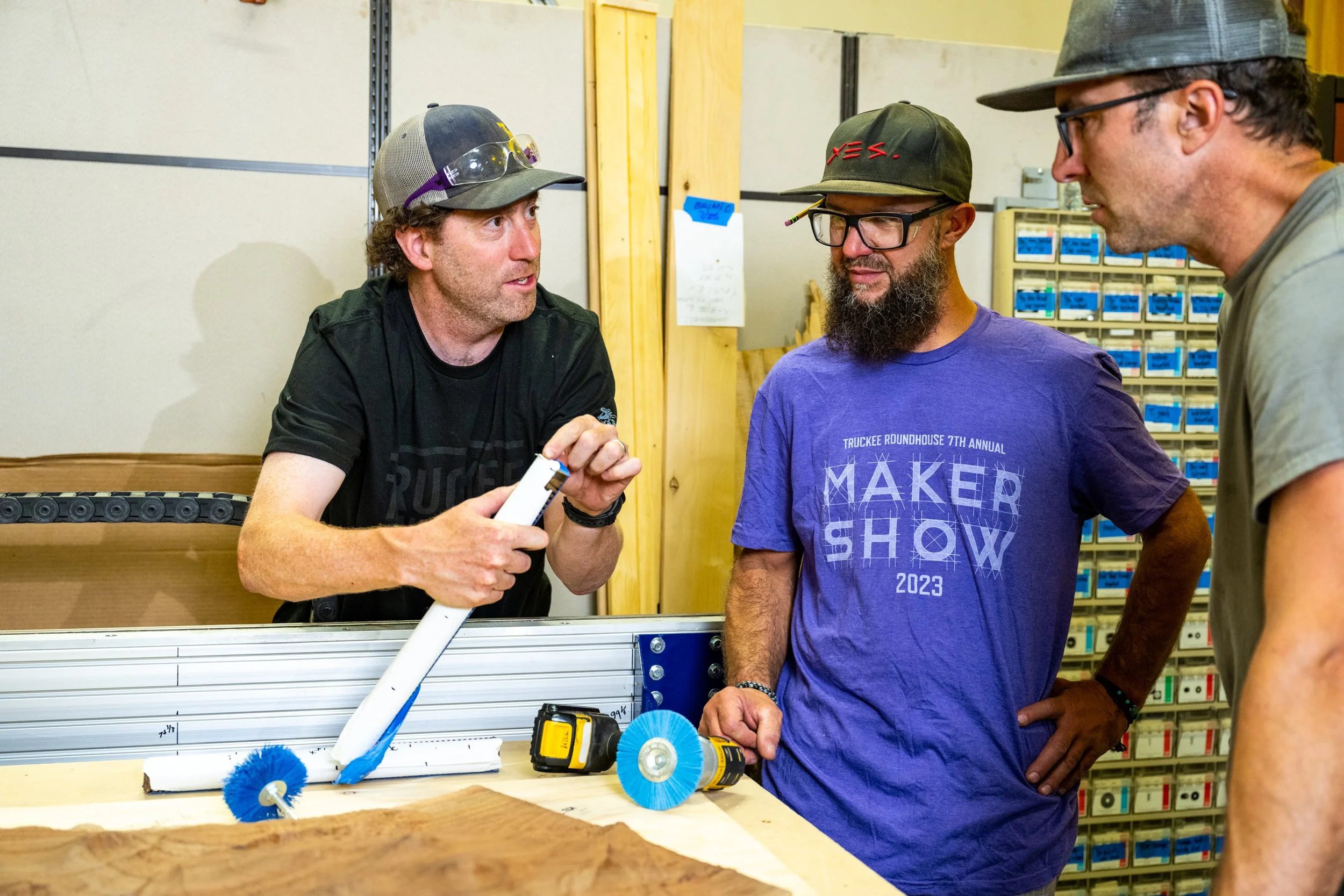 Three men are having a discussion in what looks like a workshop or maker space. One man is explaining something while holding a cylindrical object, with tools and a piece of hardware on the table in front of them. The background has organized storage bins and wooden supplies.