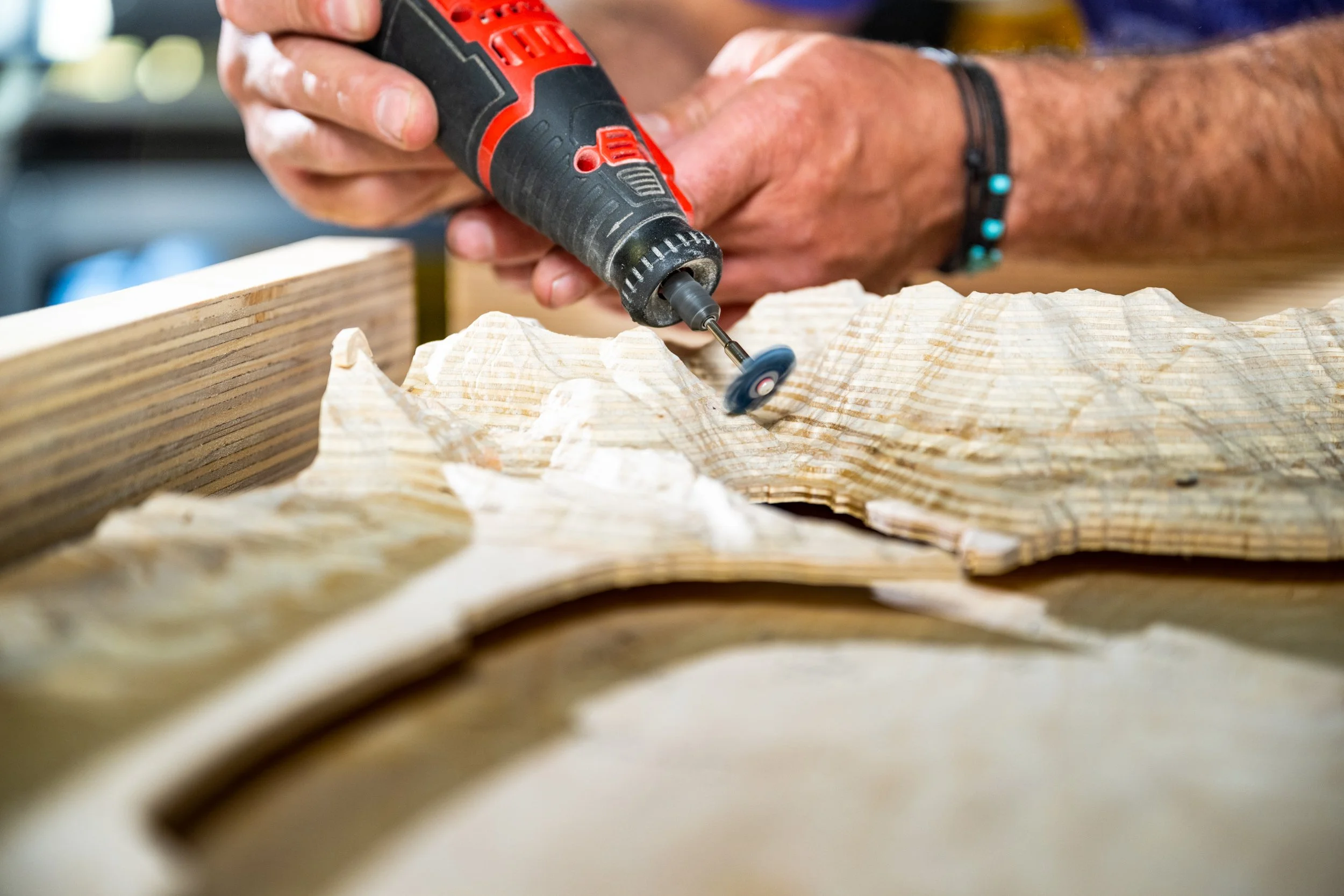 A person using a rotary tool to carve wood on a piece of wood. The person is wearing a bracelet, and the woodworking workspace appears professional.